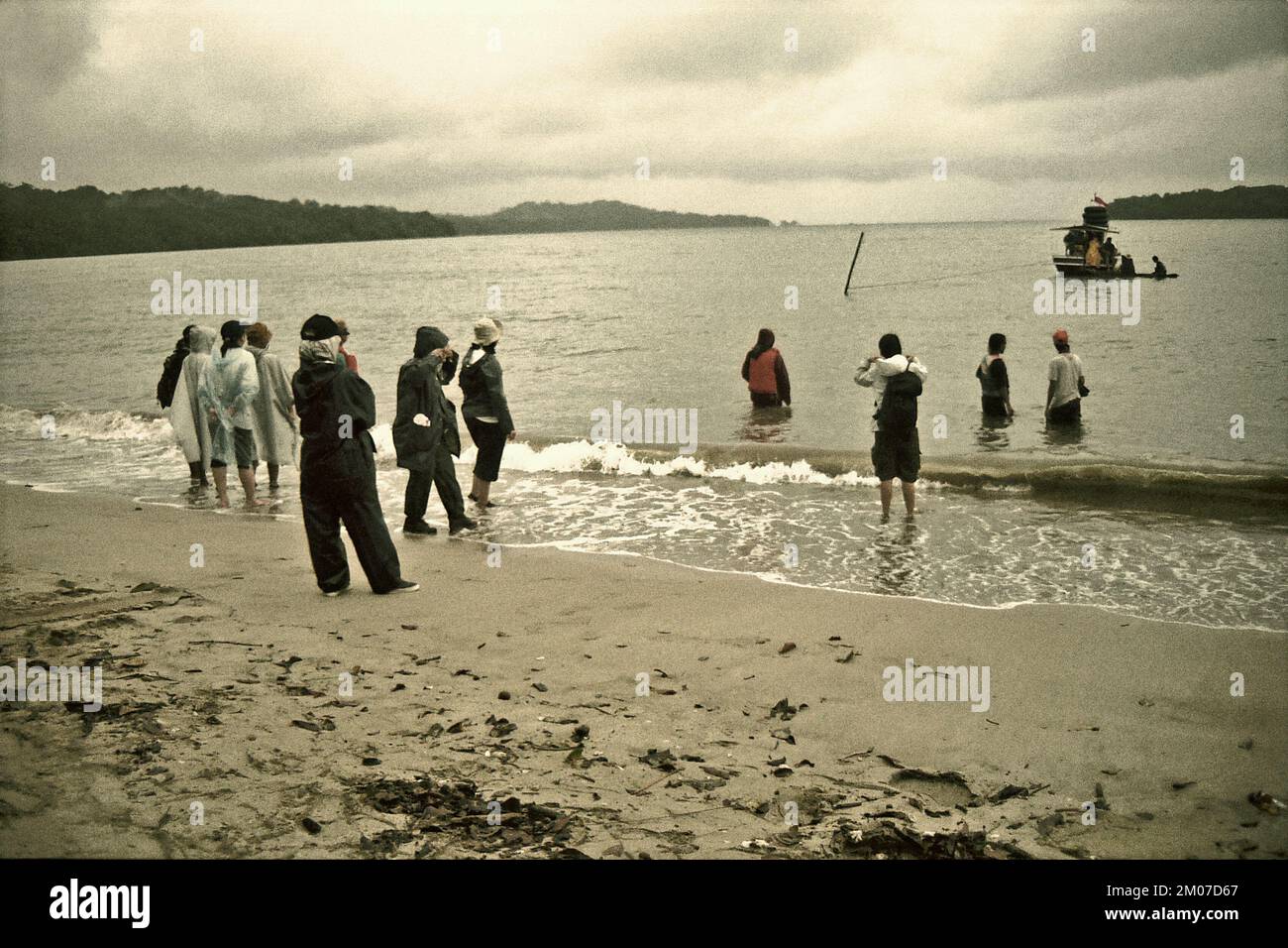 Banten, Indonésie. 5th décembre 2022. Les visiteurs se tenant sur une plage de sable, comme ils attendent d'être transportés à un bateau qui flotte sur l'eau côtière au large de la plage de Cidaon lors d'une journée de pluie lors d'une excursion écotouristique 2003 dans le parc national d'Ujung Kulon, Pandeglang, Banten, Indonésie. Le Rapport économique 2022 de l'APEC (coopération économique Asie-Pacifique), publié lors de la réunion ministérielle annuelle de l'APEC et de sa réunion des chefs de file économiques à Bangkok le mois dernier (novembre 2022), a déclaré que les membres de l'APEC sont confrontés aux défis de la durabilité. Banque D'Images