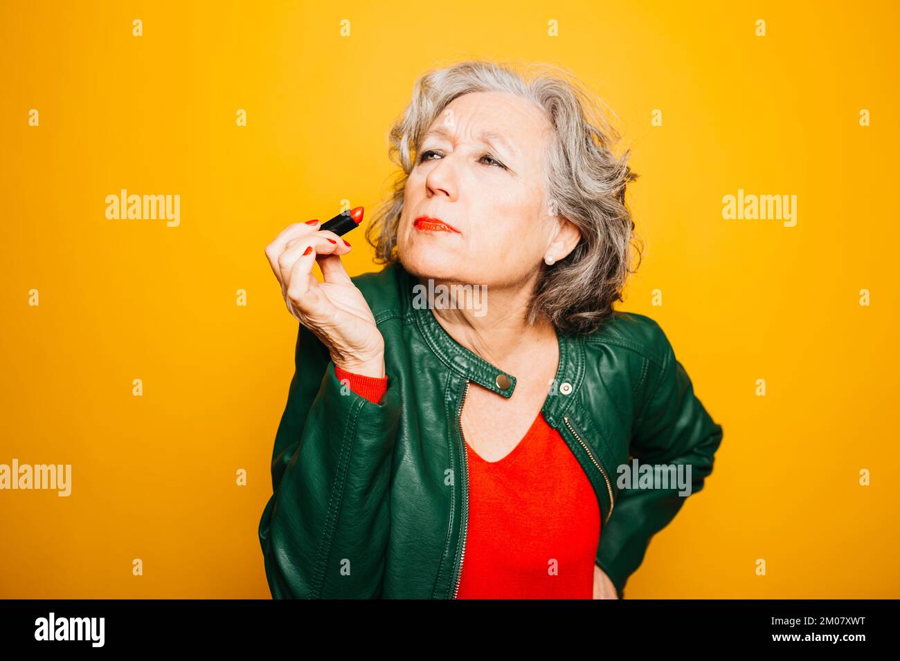 Portrait d'une femme âgée appliquant un rouge à lèvres sur un fond jaune Banque D'Images Portrait d'une femme âgée appliquant un rouge à lèvres sur un fond jaune Banque D'Images