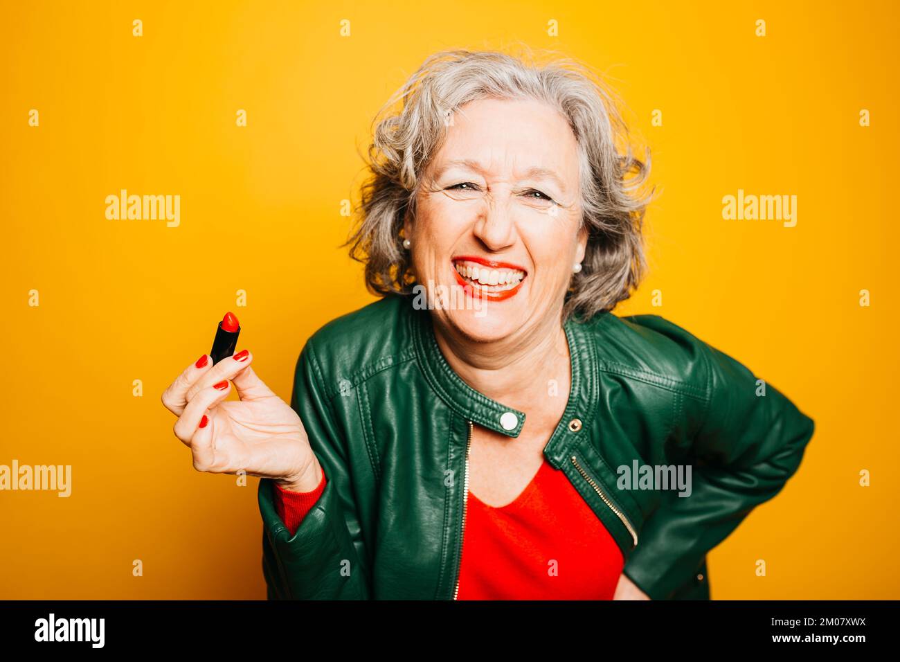 Portrait d'une femme âgée appliquant un rouge à lèvres sur un fond jaune Banque D'Images Portrait d'une femme âgée appliquant un rouge à lèvres sur un fond jaune Banque D'Images