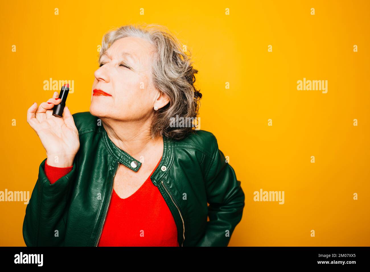 Portrait d'une femme âgée appliquant un rouge à lèvres sur un fond jaune Banque D'Images Portrait d'une femme âgée appliquant un rouge à lèvres sur un fond jaune Banque D'Images