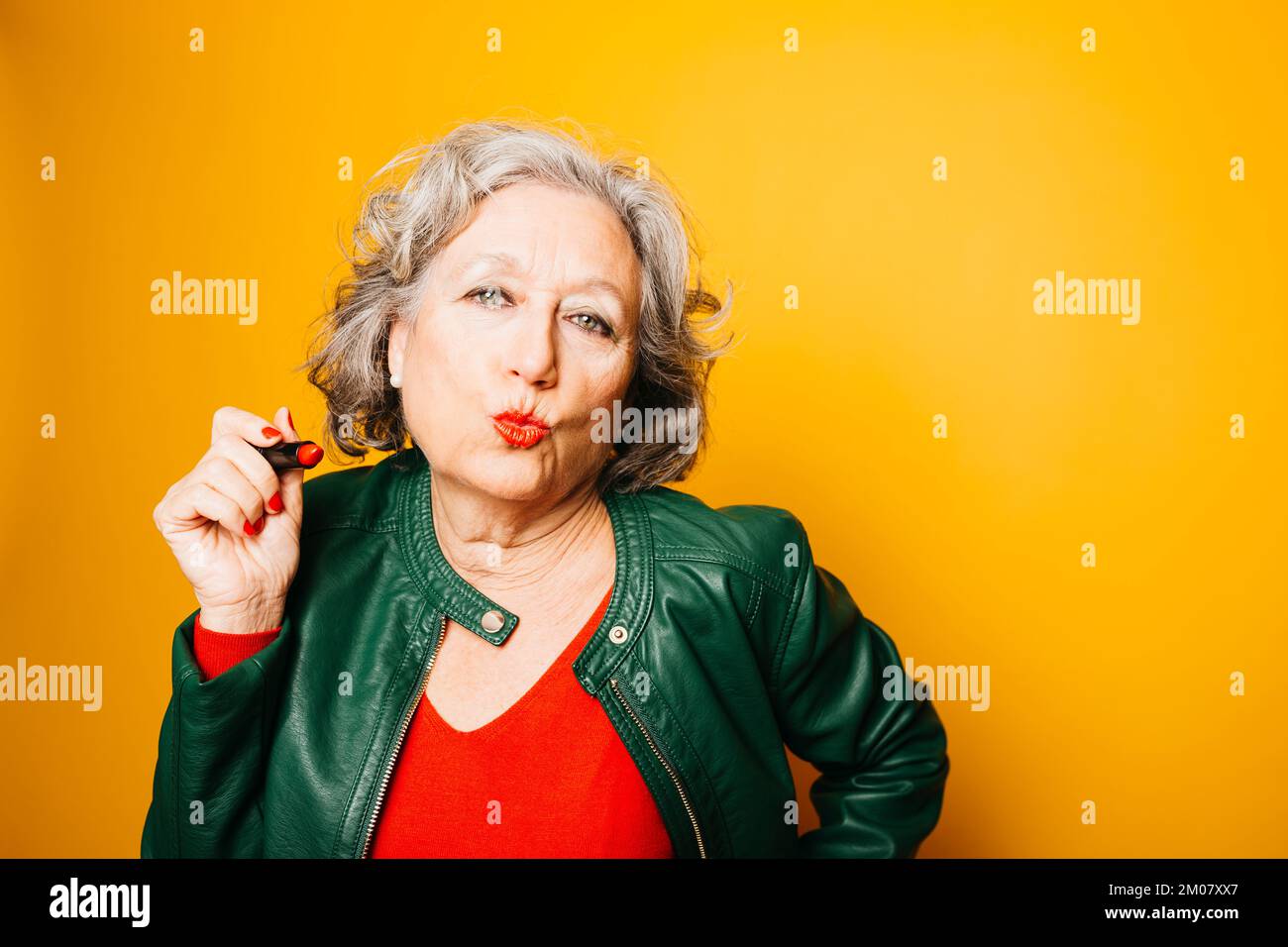 Portrait d'une femme âgée appliquant un rouge à lèvres sur un fond jaune Banque D'Images Portrait d'une femme âgée appliquant un rouge à lèvres sur un fond jaune Banque D'Images