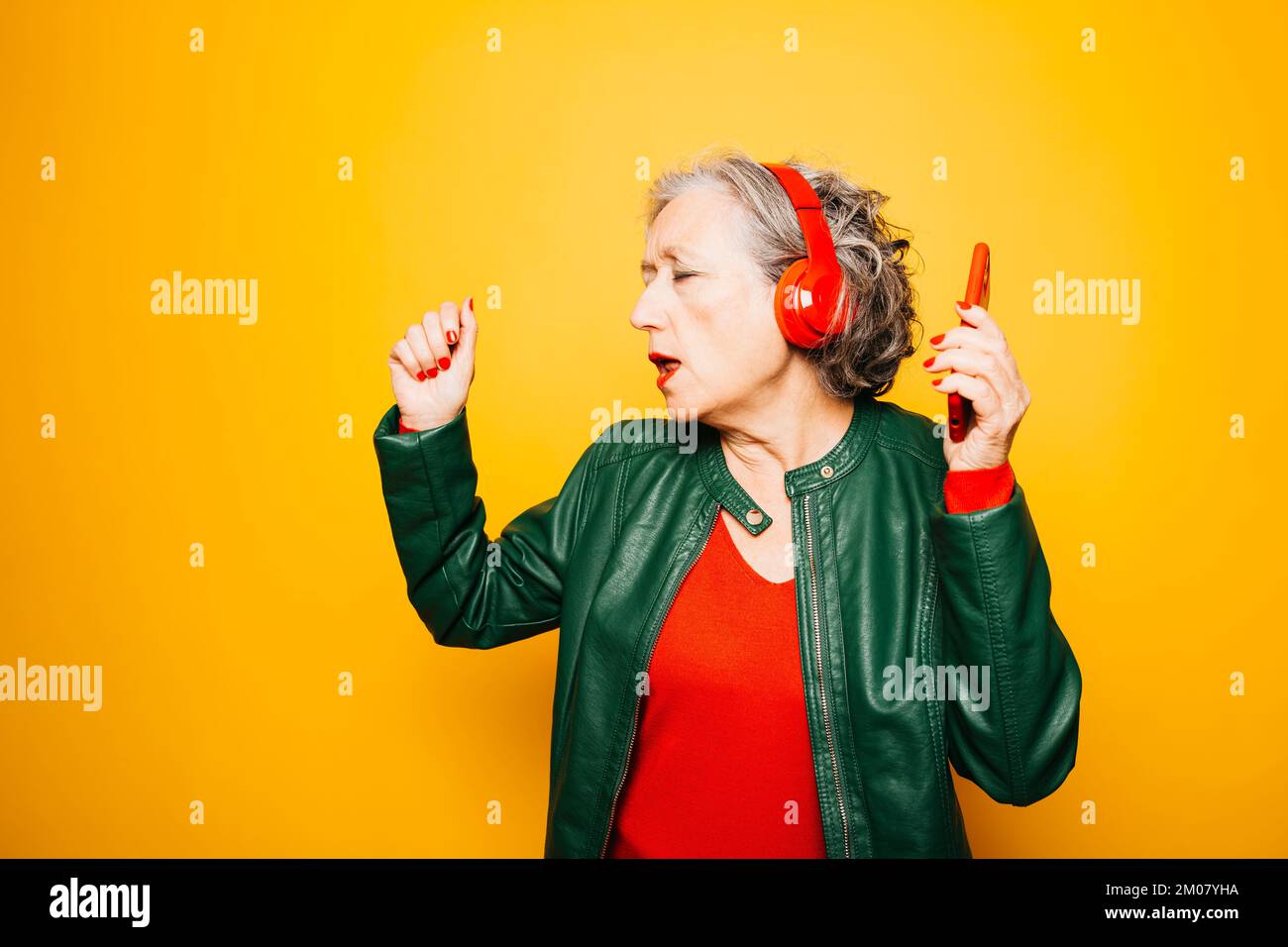 Femme âgée avec casque rouge et smartphone rouge, dansant sur fond jaune Banque D'Images
