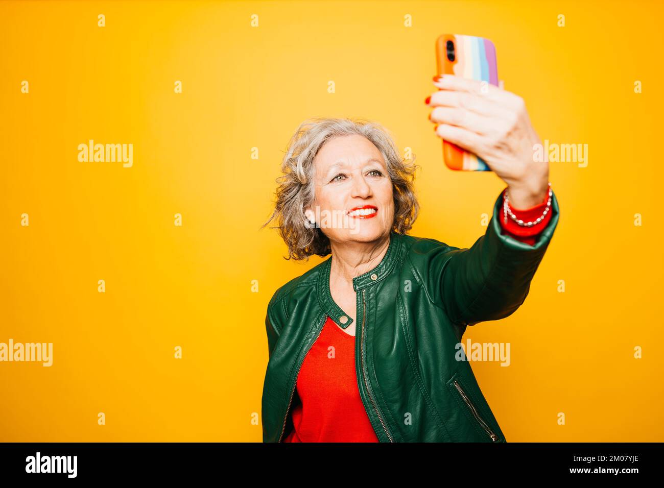 Femme âgée prenant un selfie avec un smartphone avec le drapeau arc-en-ciel sur l'étui, sur un fond jaune Banque D'Images