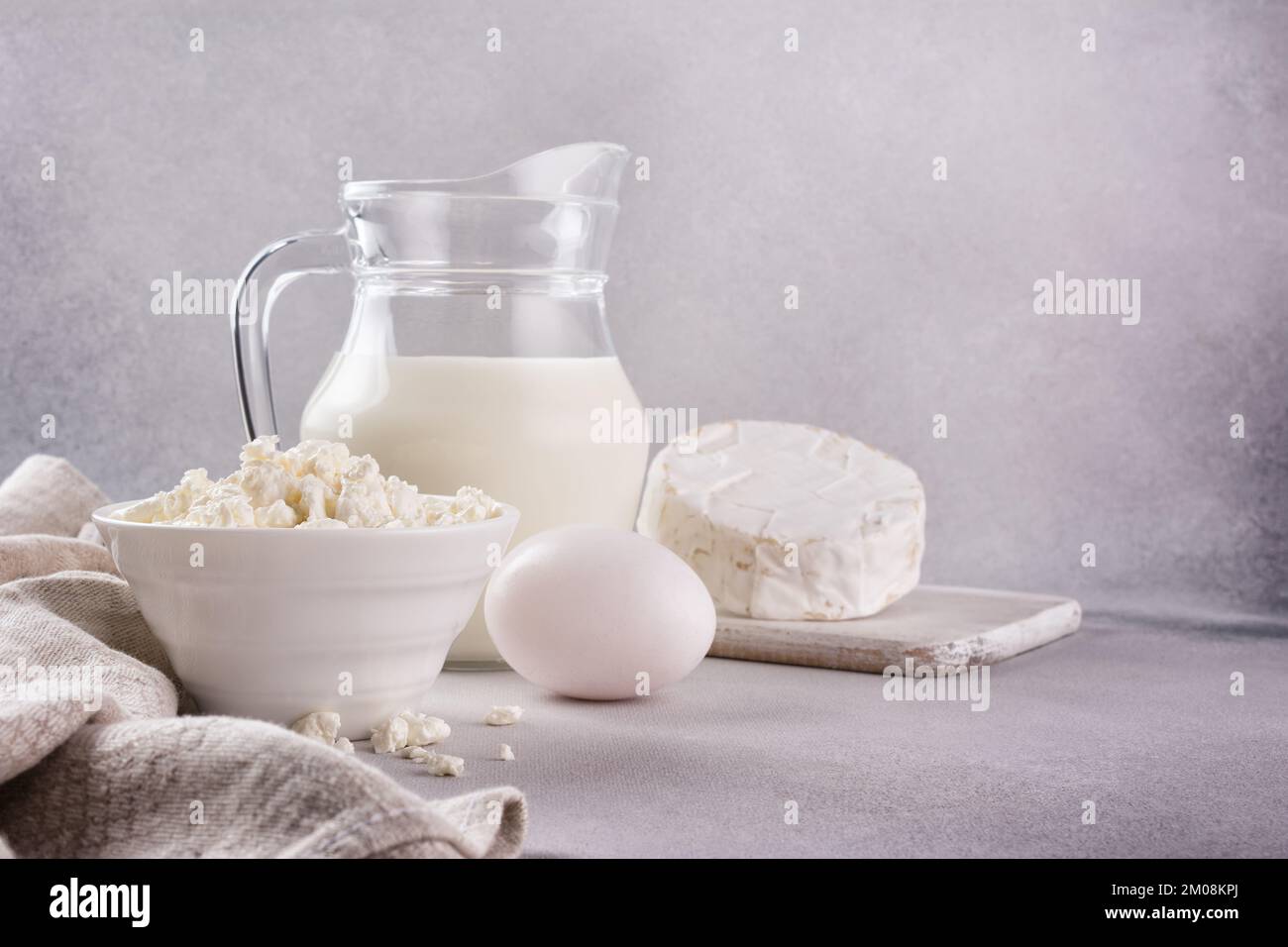 Vue de dessus du fromage cottage dans un bol blanc avec un pot à lait en verre, des œufs de poulet et du camembert comme ingrédient pour un repas savoureux sur fond de béton gris Banque D'Images