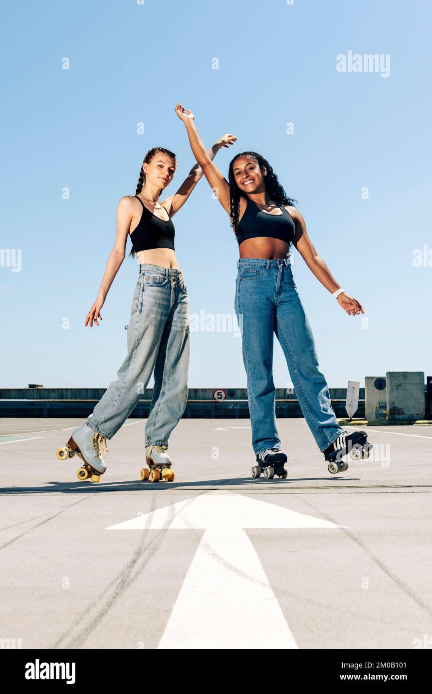 Deux filles patinent sur un parking extérieur au soleil avec leurs bras levés. Banque D'Images