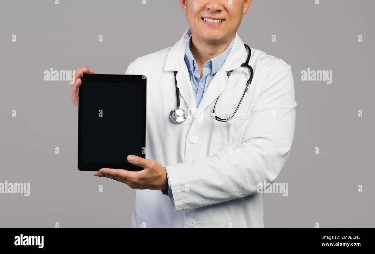 Un homme japonais souriant thérapeute en manteau blanc avec stéthoscope montre une tablette avec écran vide Banque D'Images