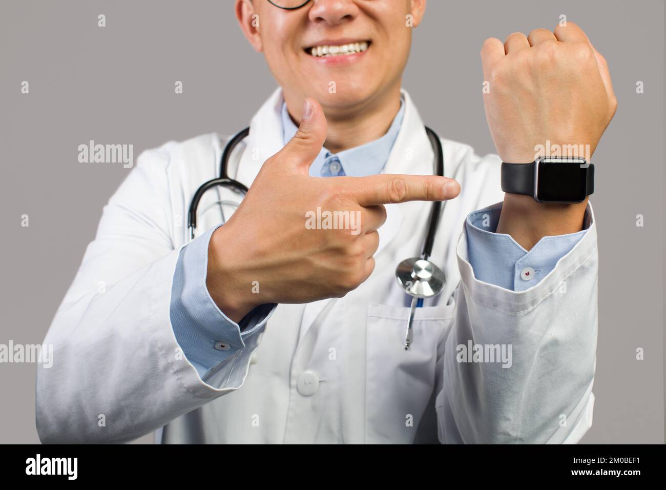 Joyeux adulte asiatique homme médecin en manteau blanc pointant le doigt à la montre avec écran vide Banque D'Images