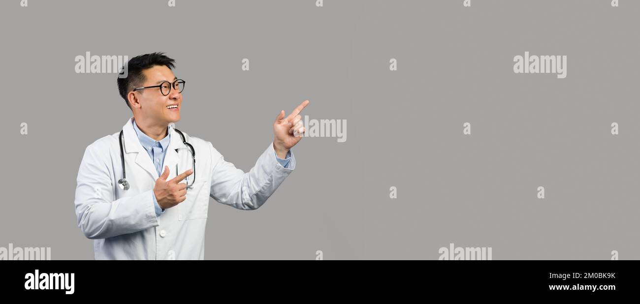 Gai homme chinois mature thérapeute en manteau blanc et des lunettes pointant les doigts sur l'espace de copie Banque D'Images