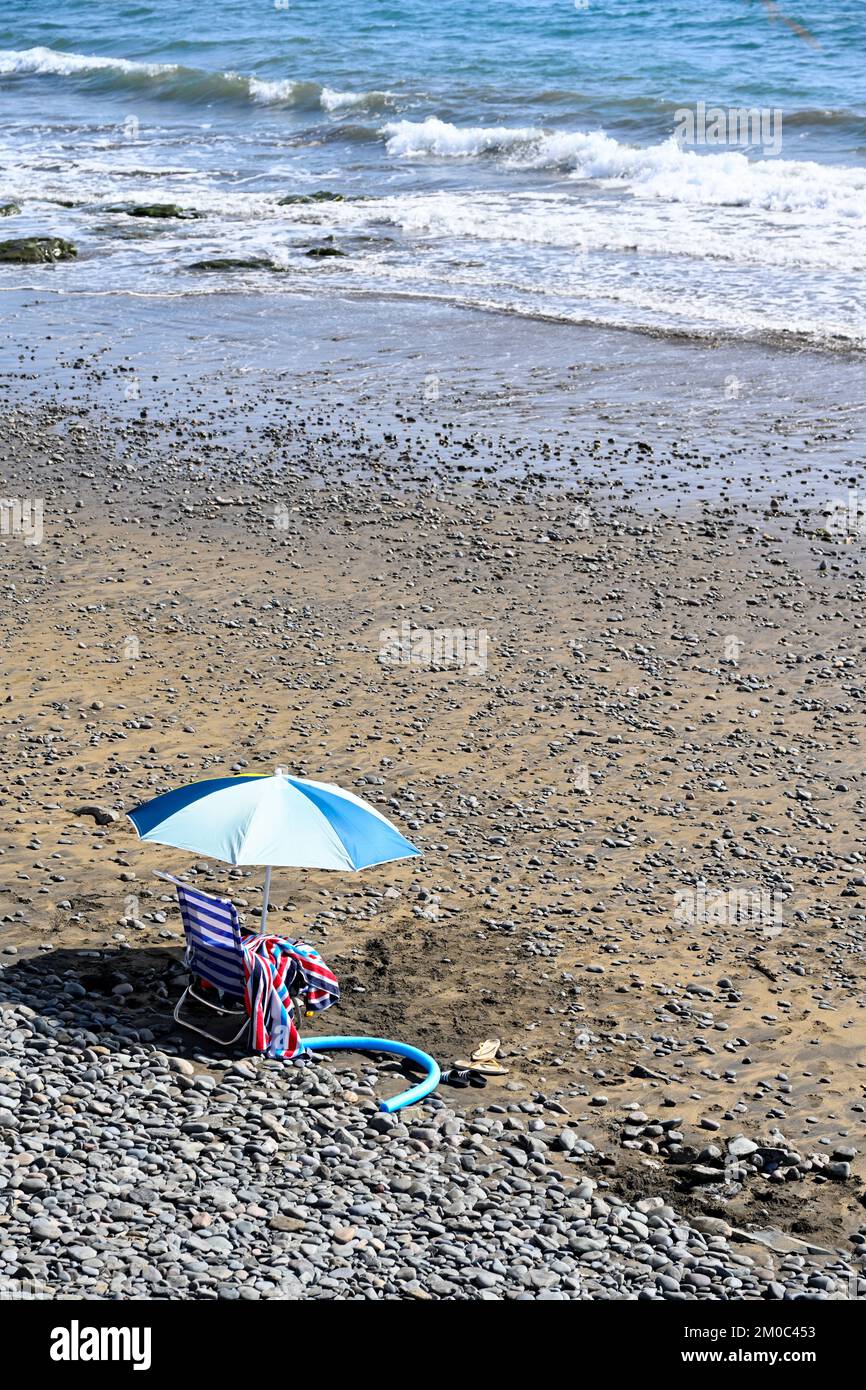 Parasol unique sur la plage de sable avec des galets assis sur une chaise pliante lisant la mer et serf calme, Playa del Águila, Gran Canaria Banque D'Images