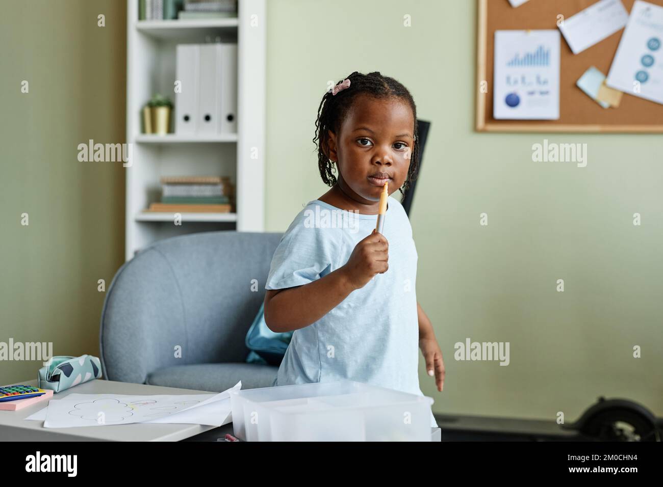 Portrait d'une jolie fille noire tenant un crayon et regardant l'appareil photo tout en dessinant des photos sur un petit bureau Banque D'Images