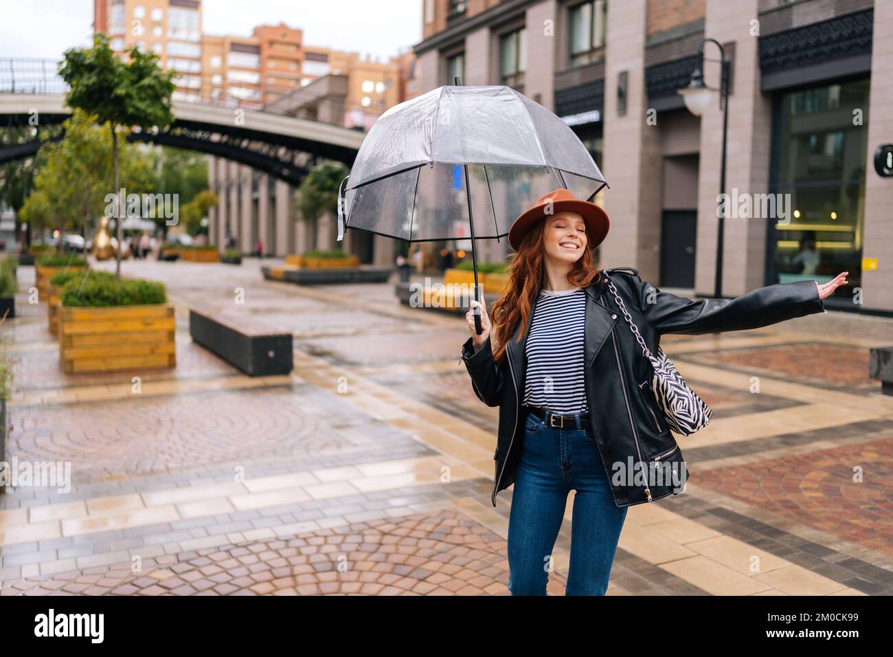 Joyeuse et excitée jeune femme dans un élégant chapeau dansant et s'amusant avec un parapluie transparent sur une belle rue de la ville, en profitant du temps pluvieux dehors Banque D'Images