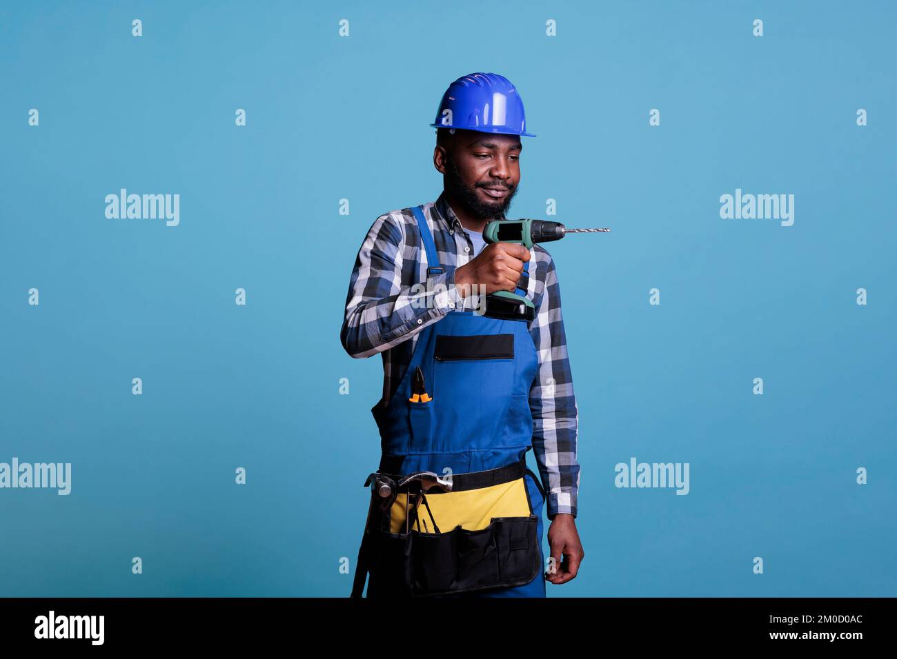 Casque de protection pour uniforme de travail afro-américain tenant une  perceuse électrique sur fond bleu. Outil de travail de maintien  professionnel de la construction utilisant une courroie avec des marteaux  Photo Stock -