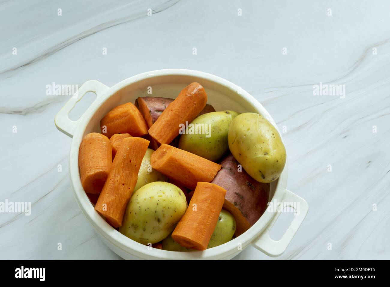 Un récipient en plastique blanc rempli de pommes de terre nettoyées et hachées, de patates douces et de carottes prêtes à cuire Banque D'Images