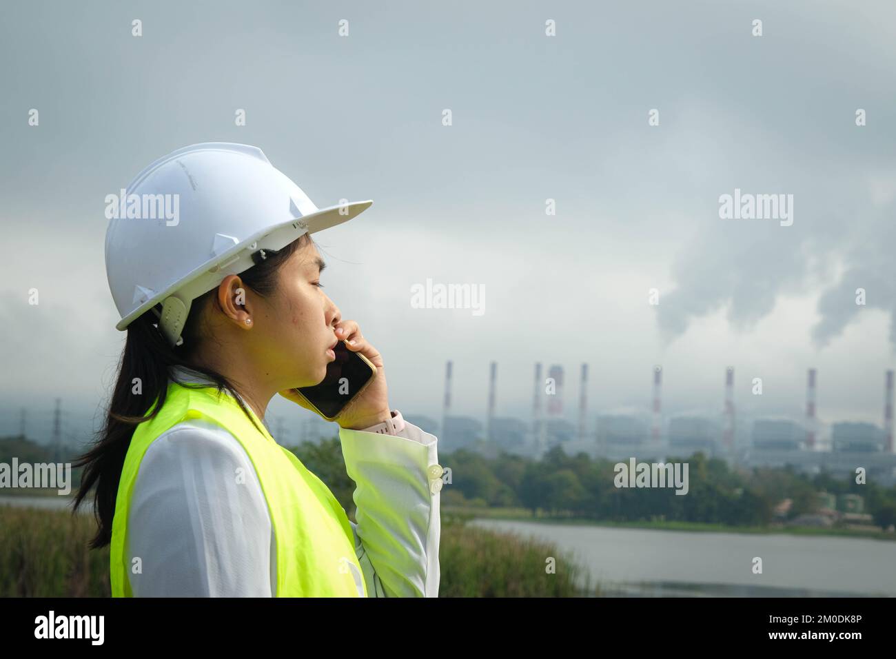 Femme chef mécanicien en gilet vert et casque parlant au téléphone à l'extérieur sur le fond de la centrale au charbon et de vapeur dans la mornin Banque D'Images