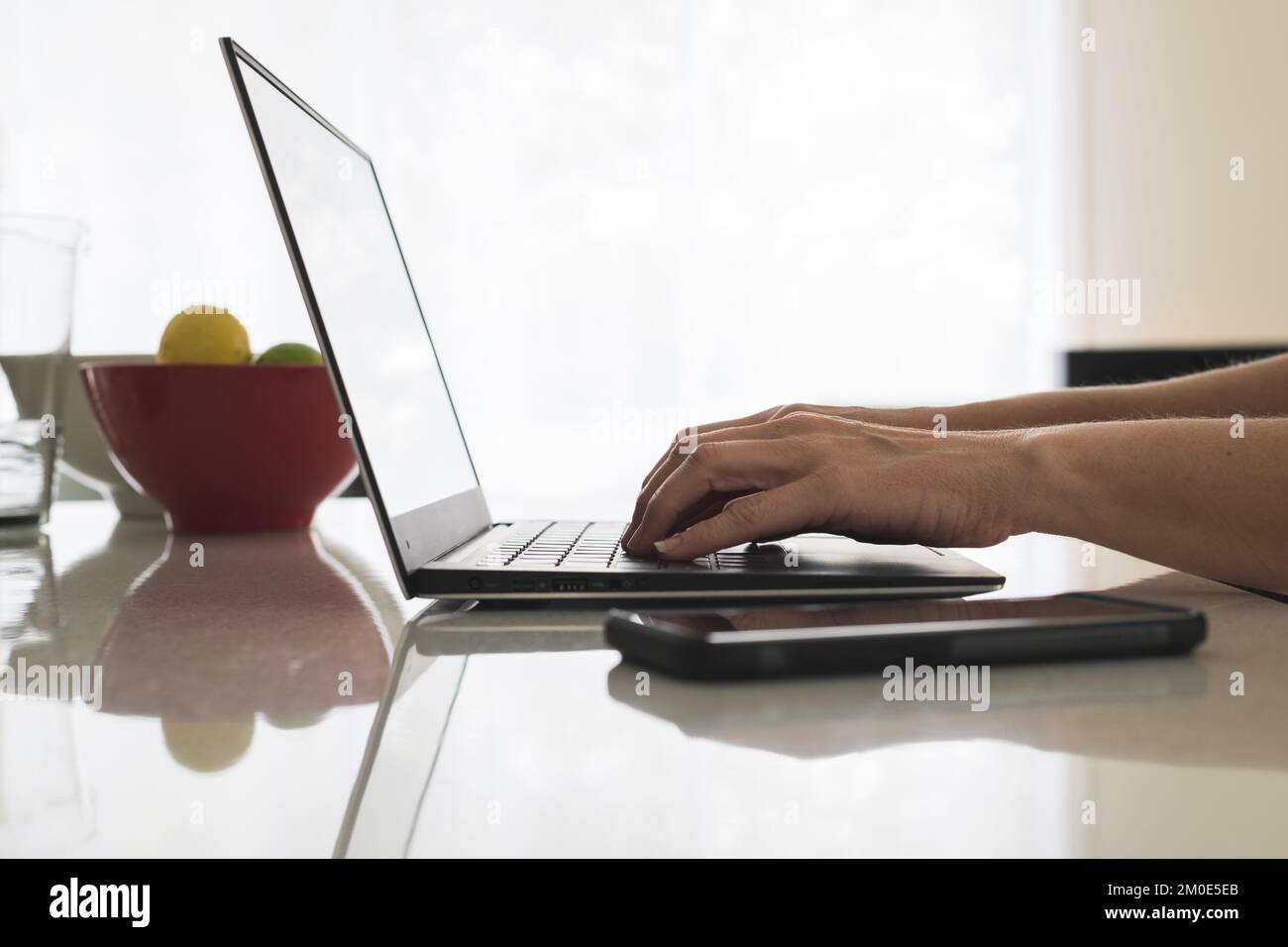 Gros plan des mains de femmes tapant sur le clavier d'un ordinateur portable moderne avec téléphone mobile à côté d'elle. Femme utilisateur d'ordinateur sur ordinateur portable au banc de cuisine. Banque D'Images
