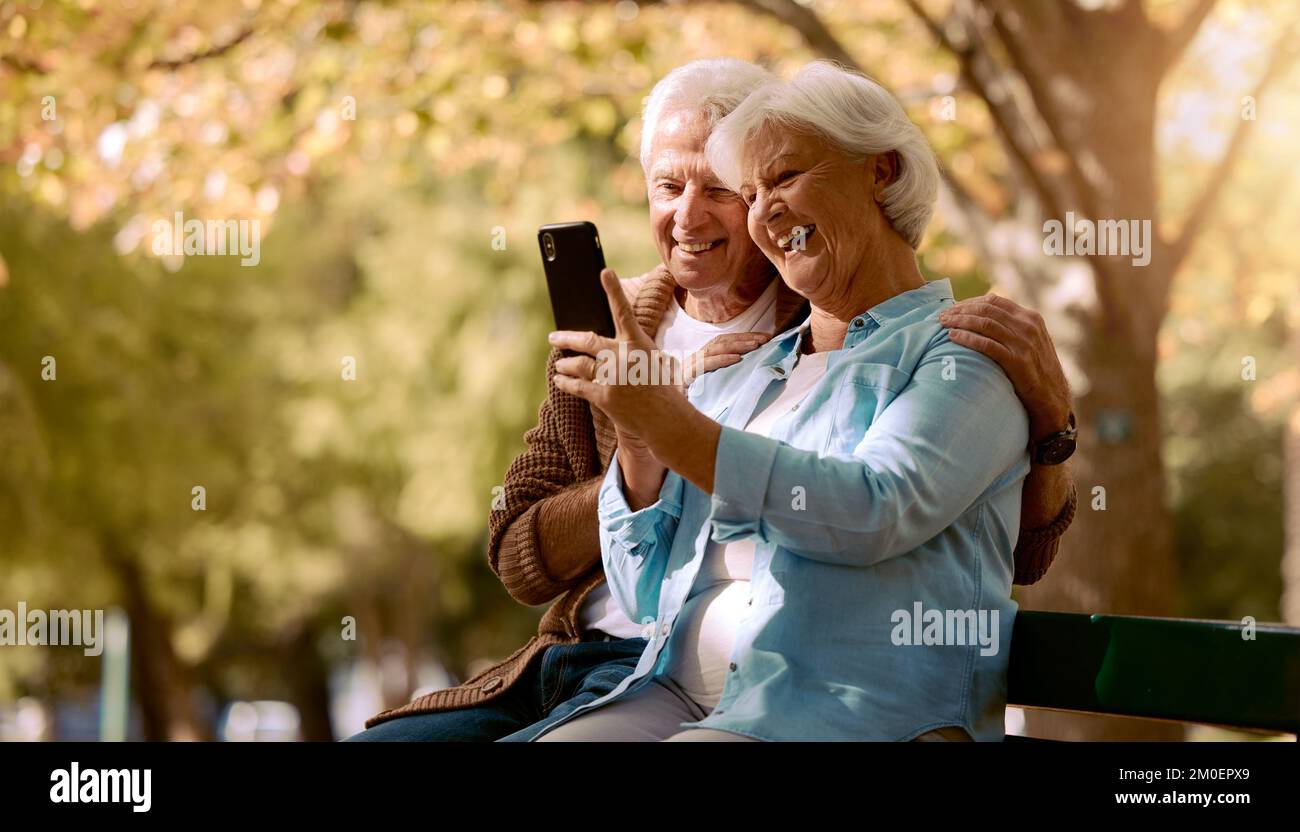 Couple senior, selfie et sourire dans le parc, heureux et arbres par un arrière-plan flou pour les médias sociaux. Homme âgé, vieille femme et smartphone sur le banc du parc Banque D'Images