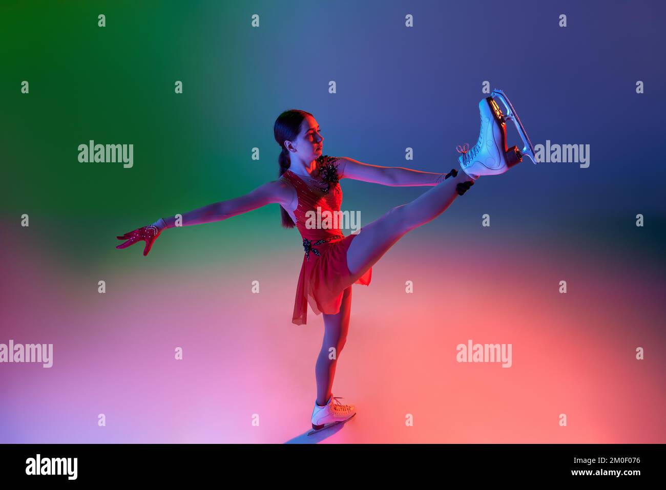 Sports d'hiver. Une patineuse féminine junior en costume de scène rouge montrant les éléments de patinage de la figure de base, les mouvements isolés sur le dégradé vert-bleu Banque D'Images