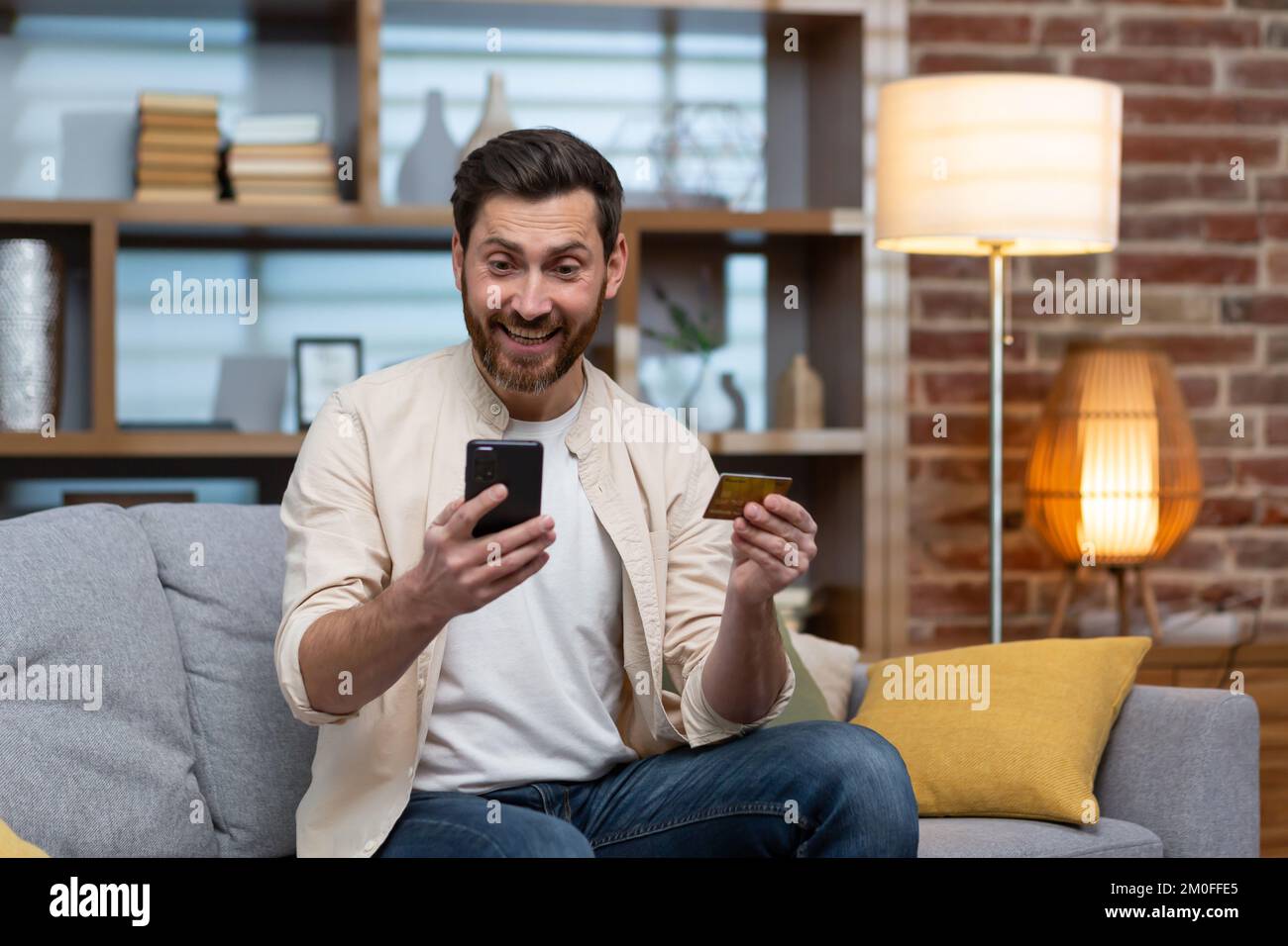 Acheteur satisfait et joyeux assis sur le canapé à la maison, homme utilisant le téléphone et la carte de crédit bancaire pour acheter des cadeaux en ligne dans la boutique en ligne. Banque D'Images