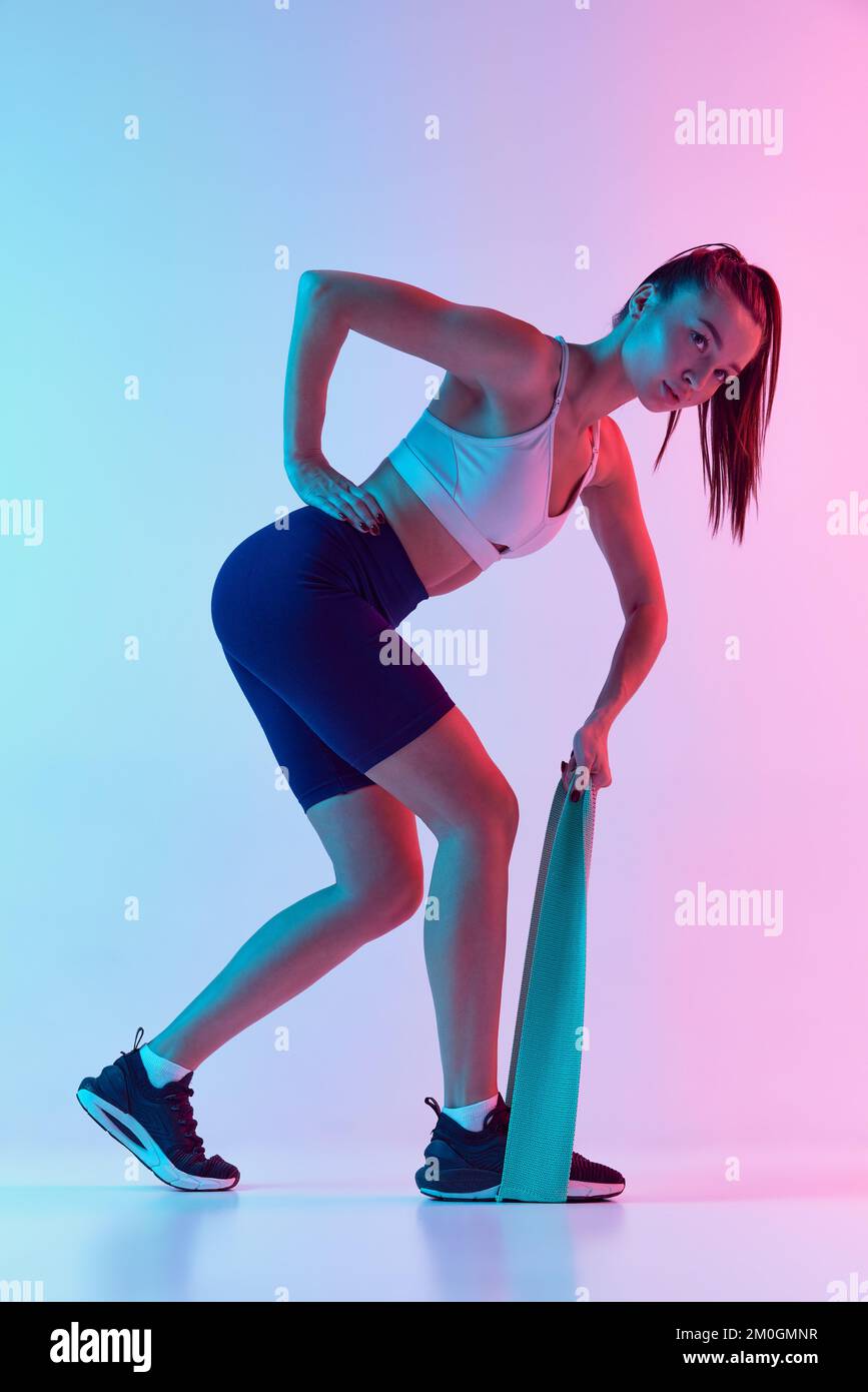 Portrait de la jeune femme sportive entraînement, faisant des exercices avec la corde d'étirement isolé sur fond bleu dégradé rose dans la lumière néon. Concept Banque D'Images