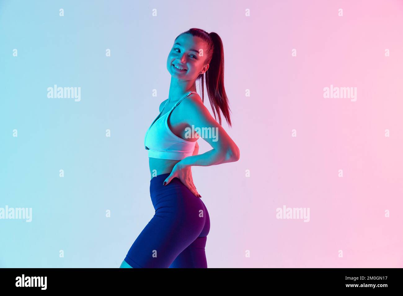 Portrait d'une jeune femme sportive dans des vêtements de sport confortables, souriant, posant isolé sur fond bleu dégradé rose dans la lumière néon. Concept de Banque D'Images