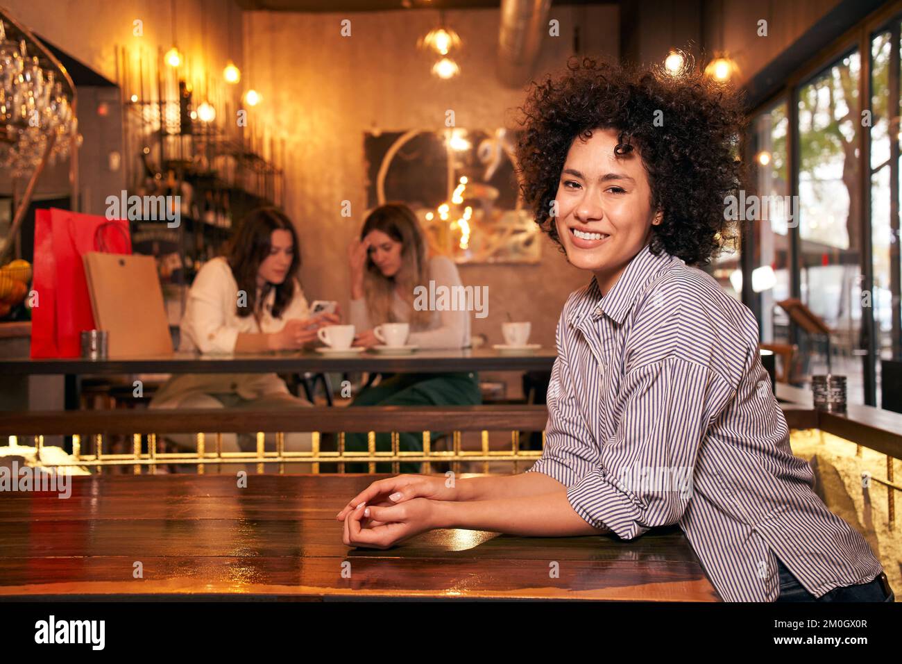 Gaie femme latine responsabilisée cheveux bouclés regardant l'appareil photo. Les copines s'amusent dans un bar Banque D'Images