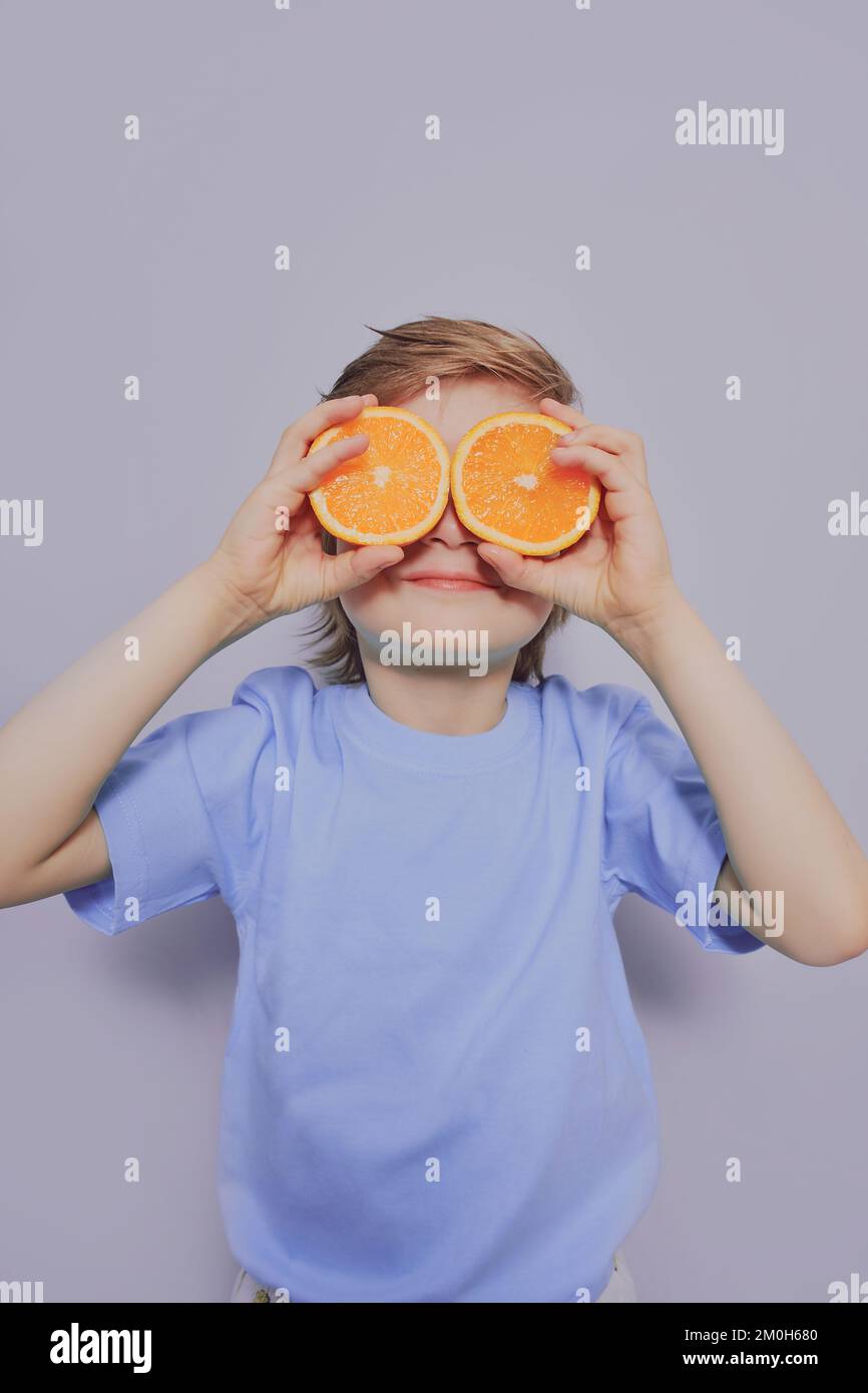 Un garçon afro-américain maudit en T-shirt blanc tient deux oranges dans ses mains et sourit sur un fond gris. Banque D'Images