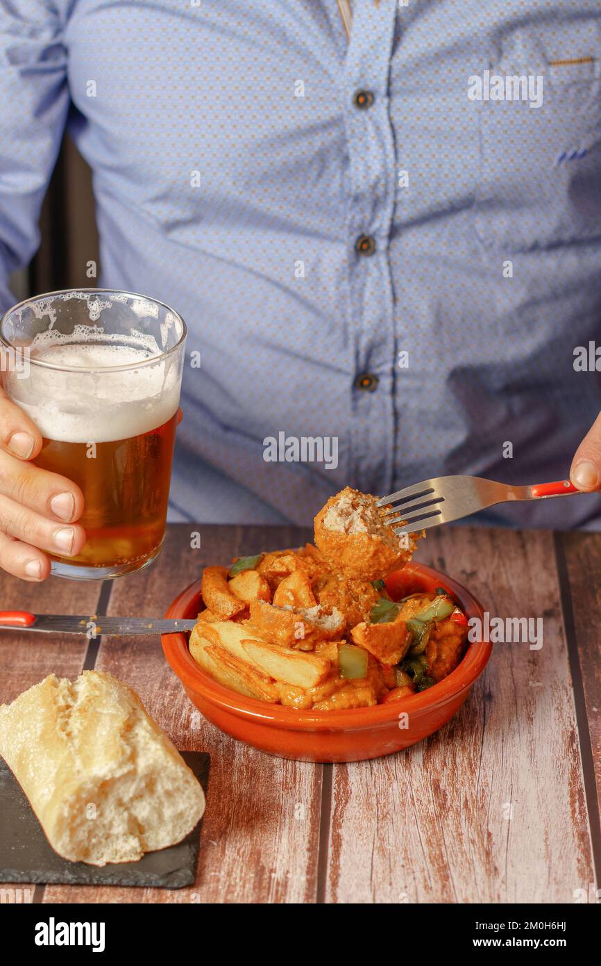 homme en chemise bleue mangeant un pot d'argile avec des boulettes de viande et des pommes de terre et une bière dans sa main Banque D'Images