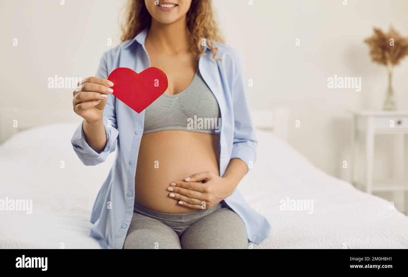 Cœur en papier rouge à la main de la femme enceinte calme et heureuse qui est dans le troisième trimestre de la grossesse. Banque D'Images