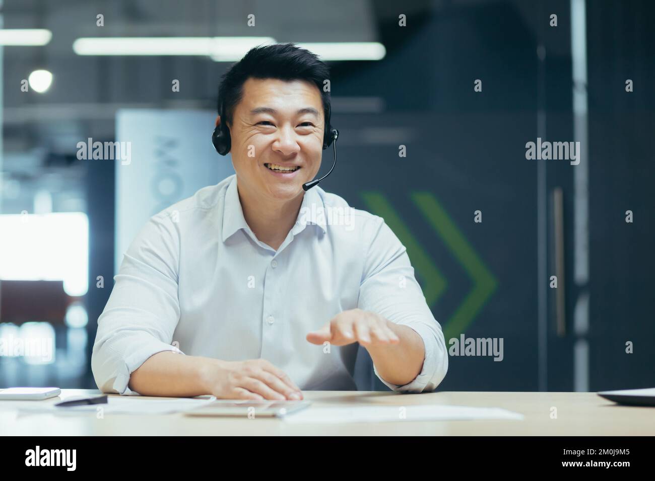 Formation en ligne. Un jeune homme asiatique souriant s'assoit et parle au bureau devant la caméra dans un casque, organise une réunion en ligne et un séminaire en ligne Banque D'Images