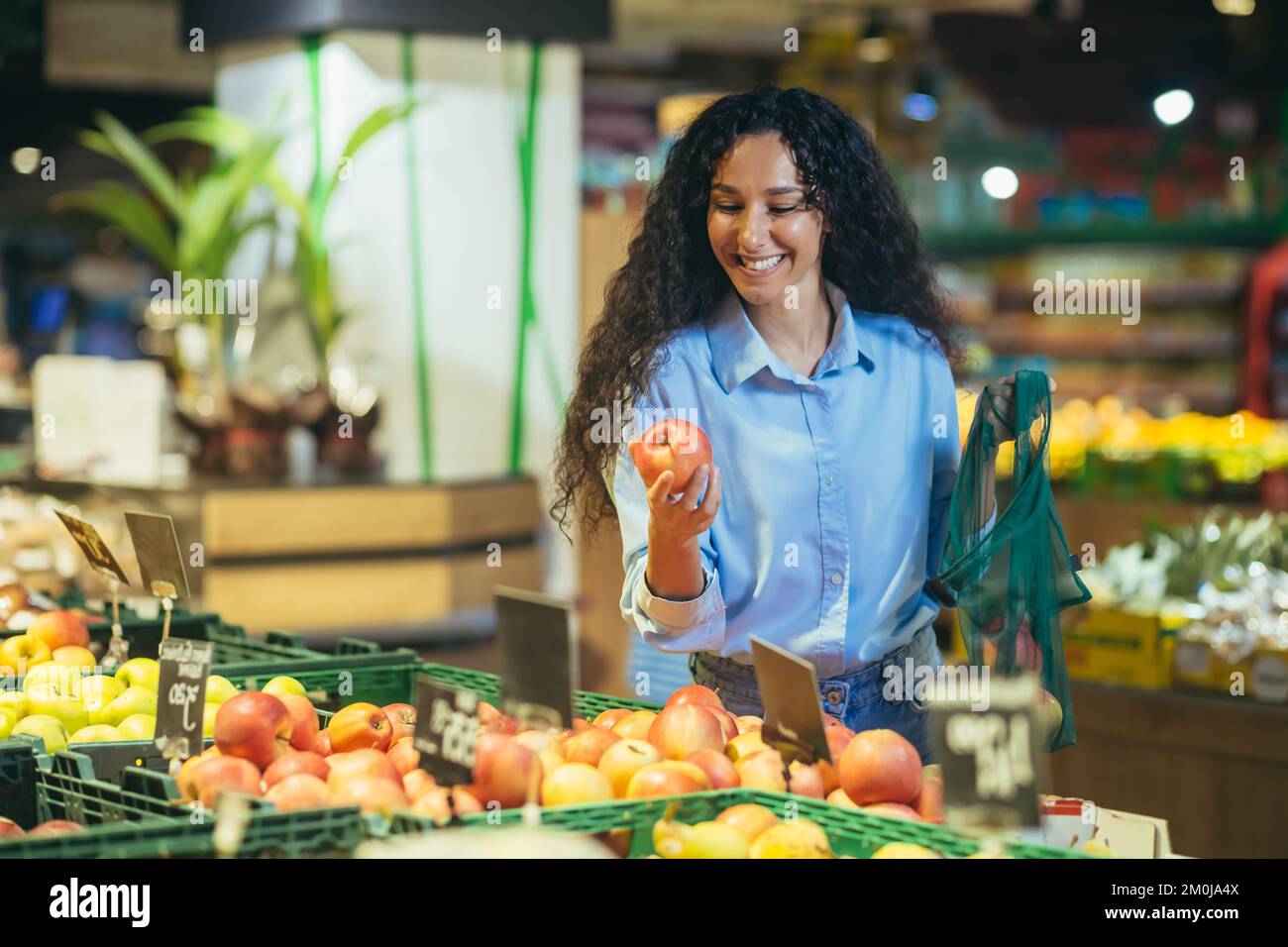 Une belle jeune femme hispanique fait du shopping dans un supermarché. Elle se tient souriante à la section des légumes et des fruits, ramasse et tient une pomme dans ses mains, la met dans un sac écologique. Banque D'Images