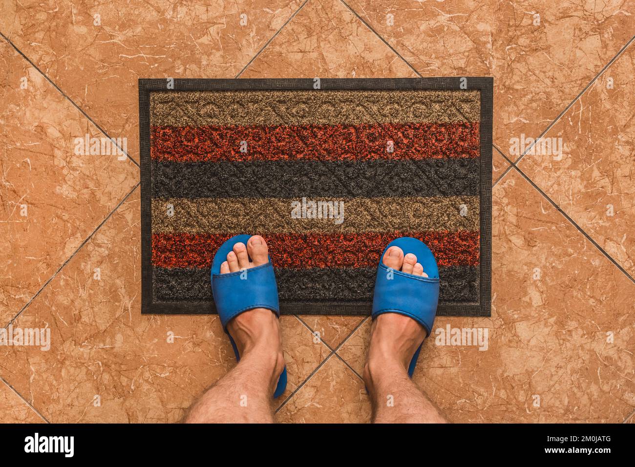 Les pieds mâles dans des pantoufles maison bleues se tiennent sur un tapis de pied sur un fond de sol carrelé brun texture, vue de dessus. Banque D'Images