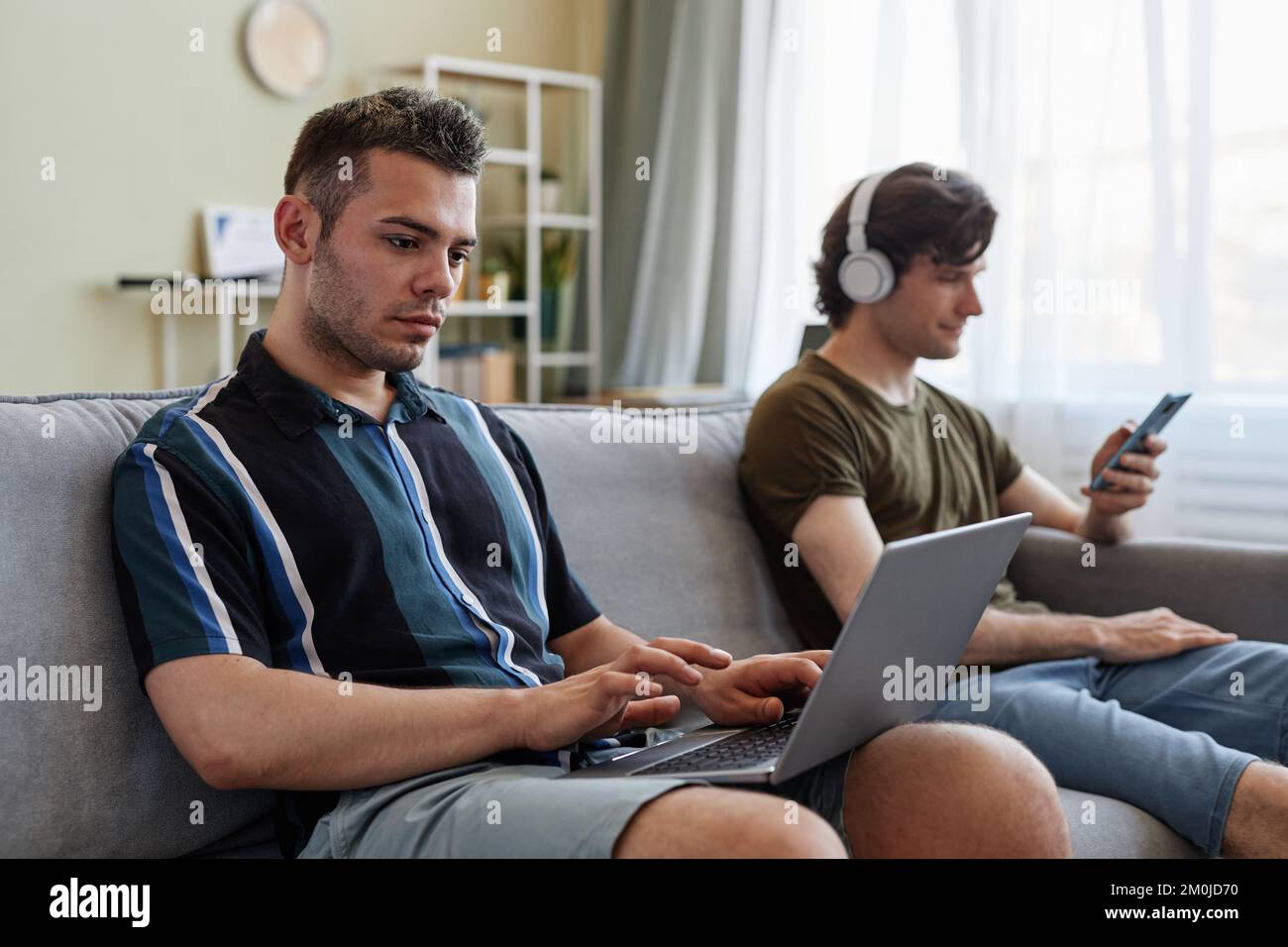 Portrait de deux jeunes hommes vivant ensemble et se reposant sur un canapé à l'aide d'ordinateurs Banque D'Images