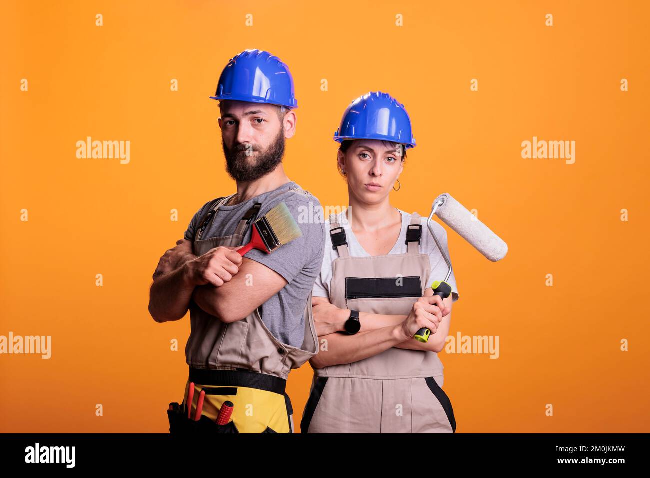 Rénovateurs professionnels posant avec des outils de peinture, debout sur fond jaune. Équipe de constructeurs qui tiennent le pinceau et le pinceau à roulettes pour travailler sur un projet de rénovation en studio. Banque D'Images