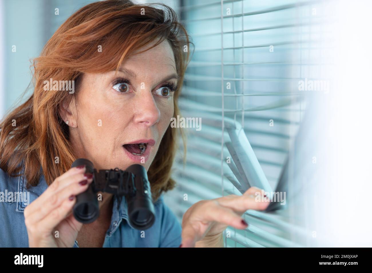 une femme adulte choquée qui espionne la fenêtre avec des jumelles Banque D'Images
