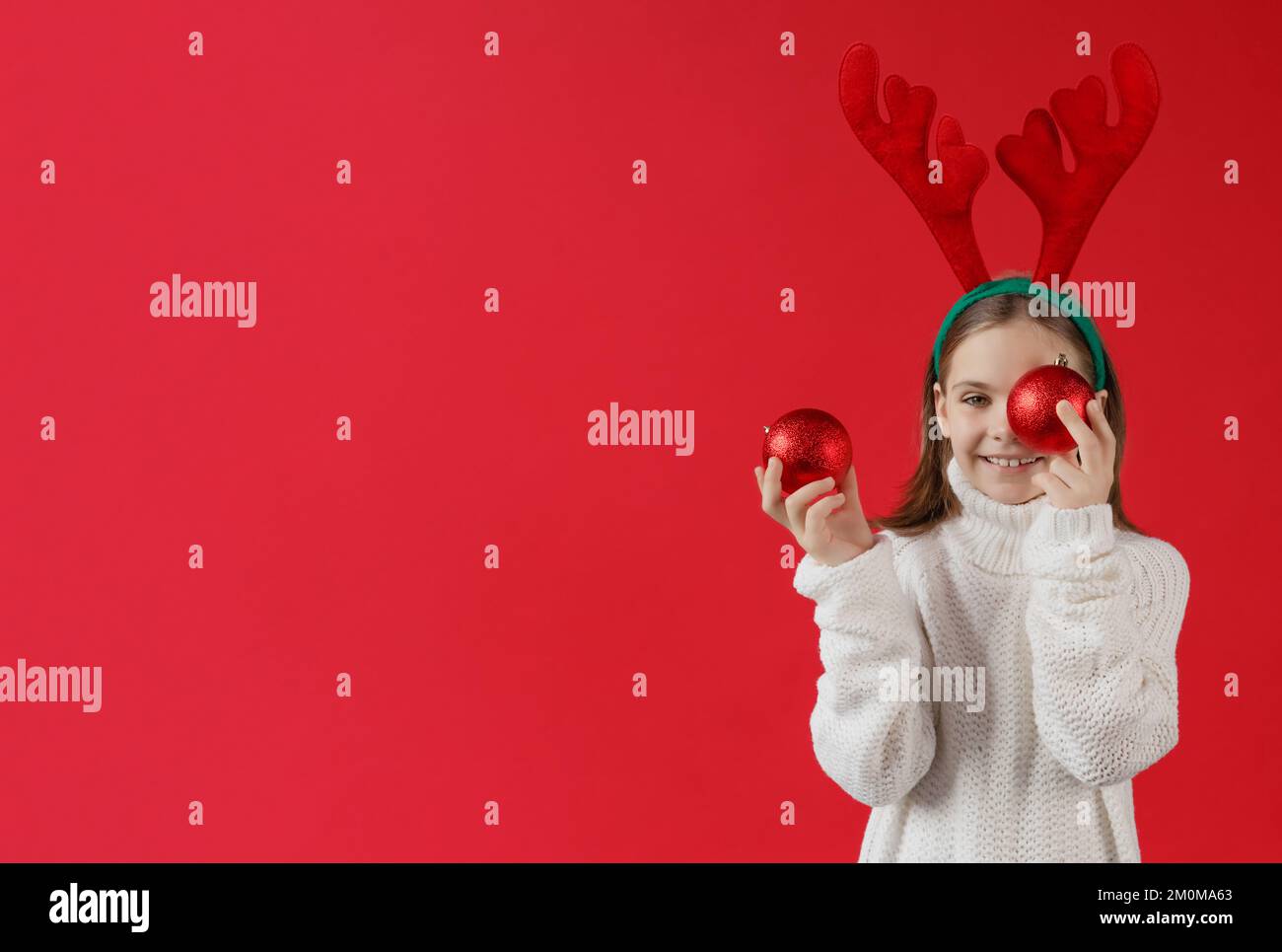 caucasienne fille avec serre-tête sous la forme de biches est debout et se cache derrière le jouet de Noël rouge sur fond rouge en chandail blanc Banque D'Images