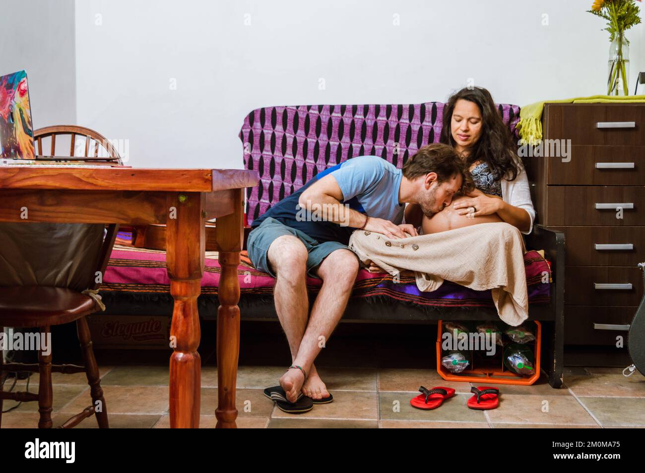 Heureux futur père Caucasien Argentin adulte avec la barbe embrasse le ventre de sa femme enceinte assis sur le canapé dans le salon, se reposant à Banque D'Images