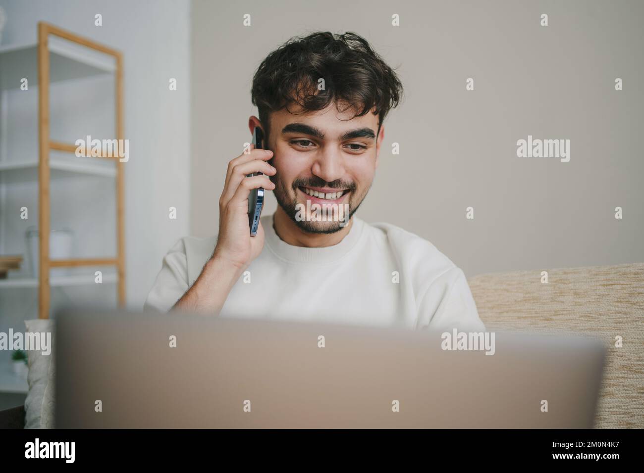 Jeune homme souriant et attrayant qui navigue sur son ordinateur portable et parle Oh téléphone mobile, tout en étant assis à la maison sur le canapé. Travail indépendant à domicile Banque D'Images