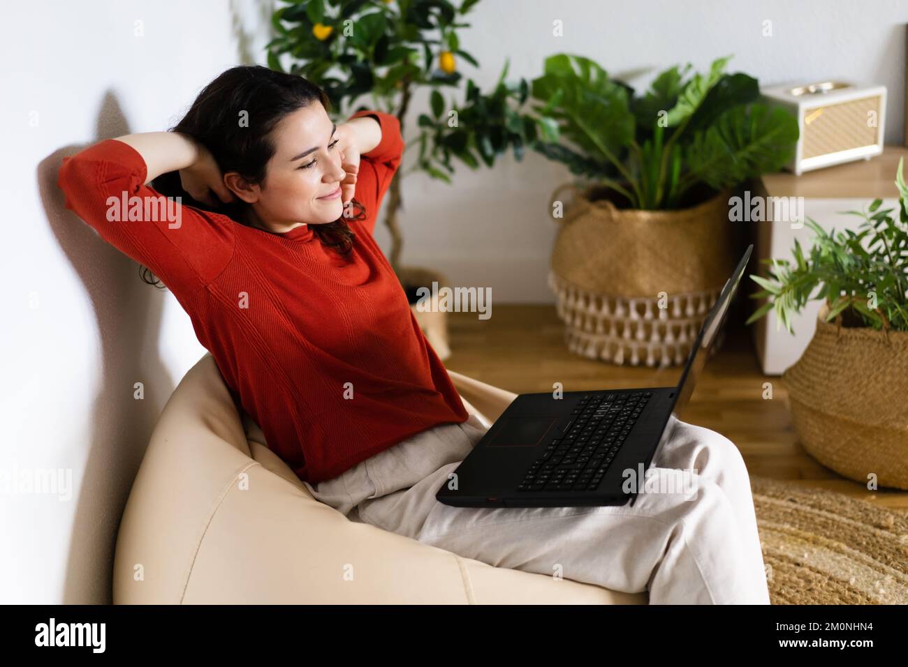 Jeune femme décontractée assise dans un fauteuil à sac avec des bras tendus à l'aide d'un ordinateur portable entouré de plantes artificielles Banque D'Images