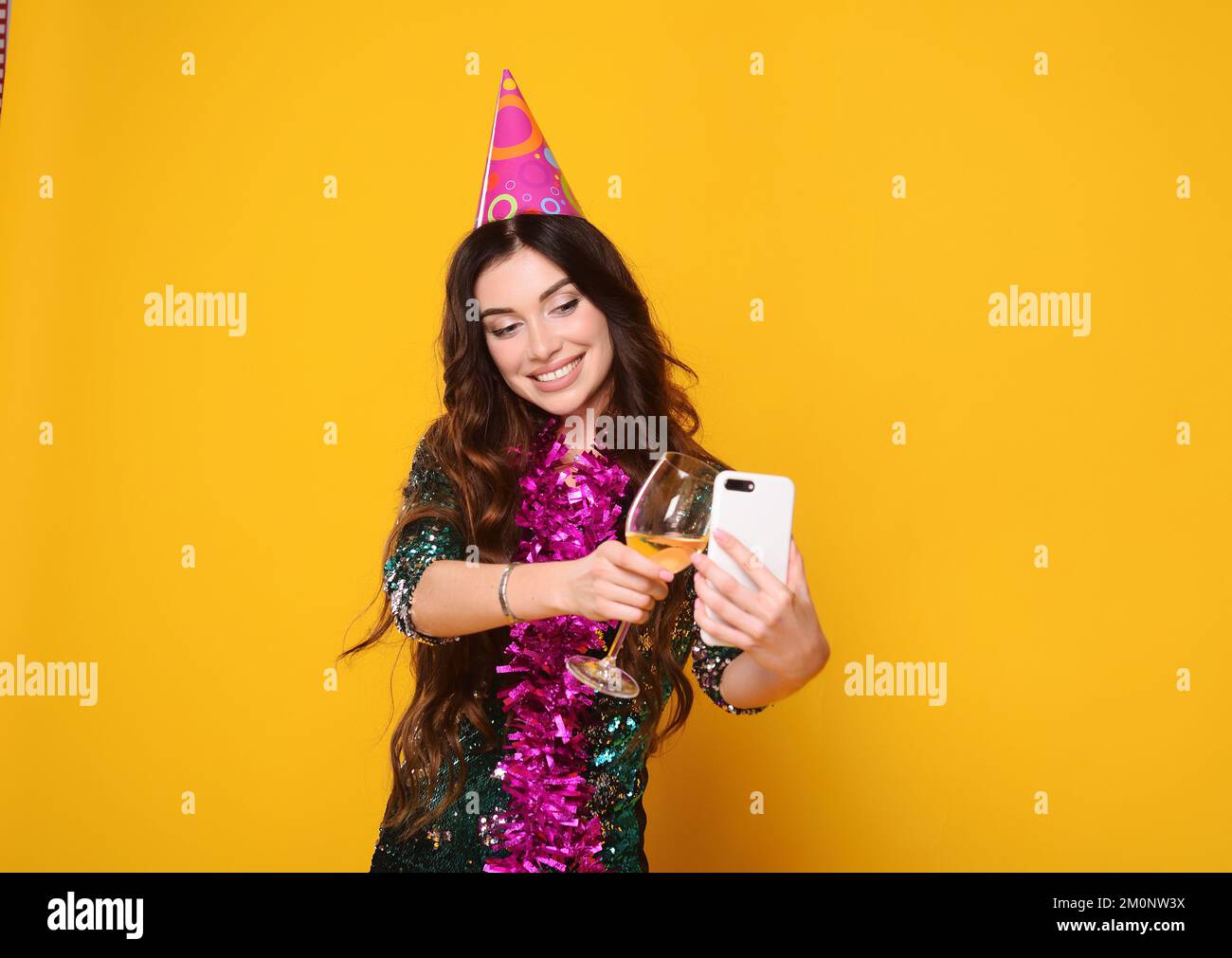 jolie femme dans un chapeau de fête rose et le boa rose tient un verre de champagne dans ses mains, sourit et tient un smartphone dans ses mains Banque D'Images