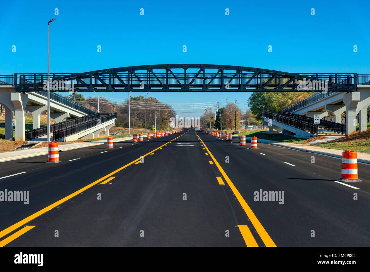 Des bandes récemment peintes et des tonneaux de circulation orange sont visibles sur une route fraîchement recouverte de noir, sous un pont piétonnier. Banque D'Images