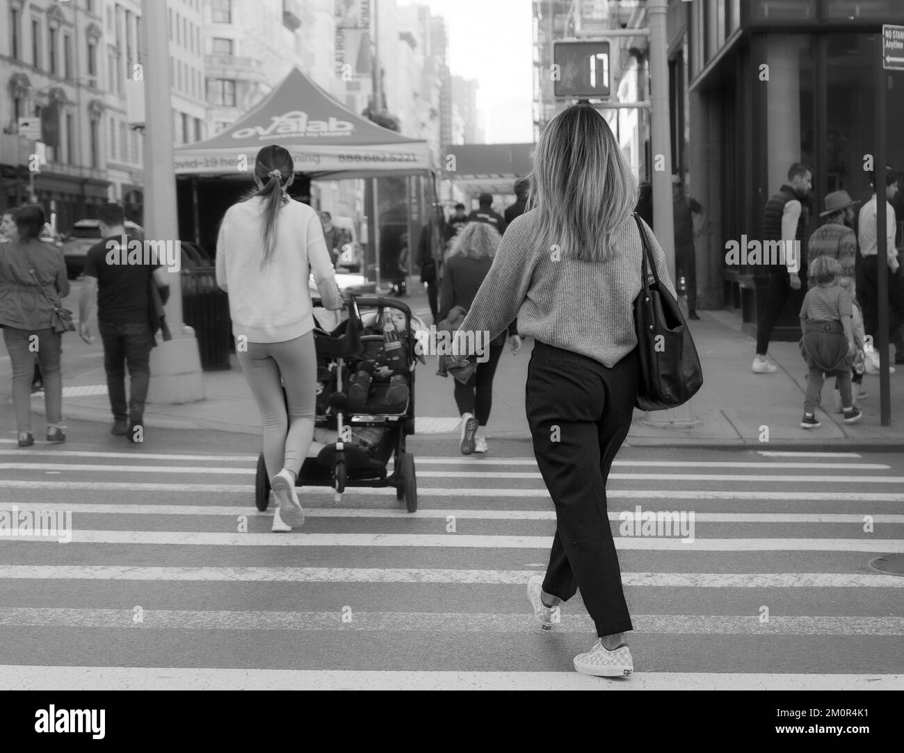 Jeune femme blonde traversant le passage de passage - photo de derrière. Photographie urbaine en noir et blanc. Jeune mère avec poussette et bébé en arrière-plan Banque D'Images