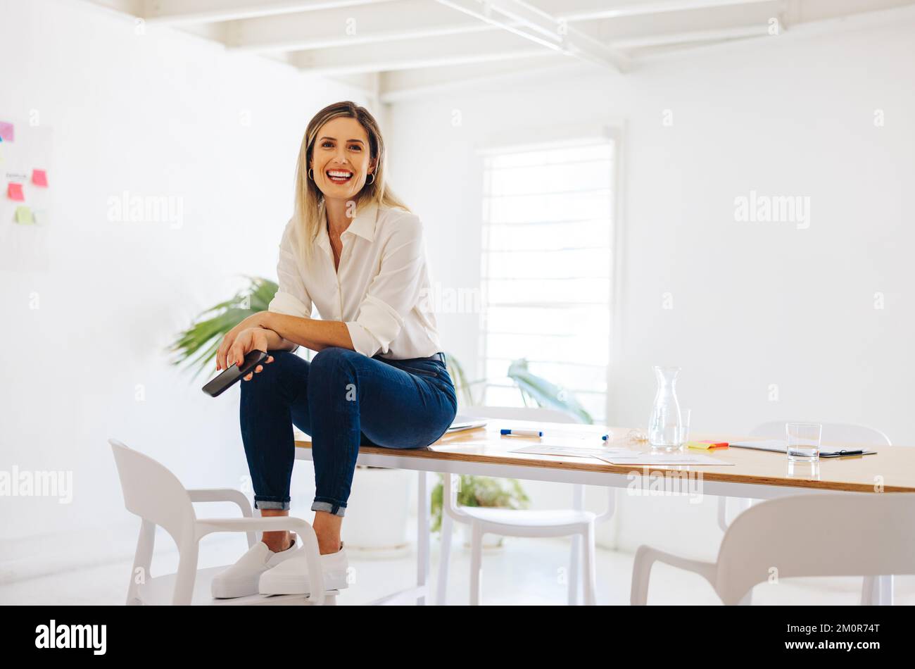 Jeune femme d'affaires souriant à la caméra tout en étant assise sur une table de conférence dans une salle de conférence moderne. Bonne jeune femme d'affaires travaillant dans un W créatif Banque D'Images