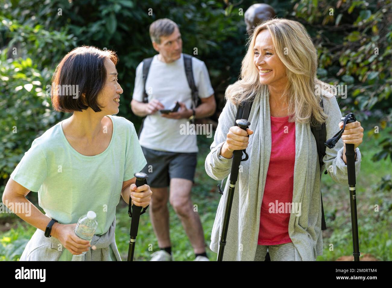 Deux dames d'âge moyen avec des bâtons de randonnée marchant dans les bois avec leurs partenaires derrière eux Banque D'Images