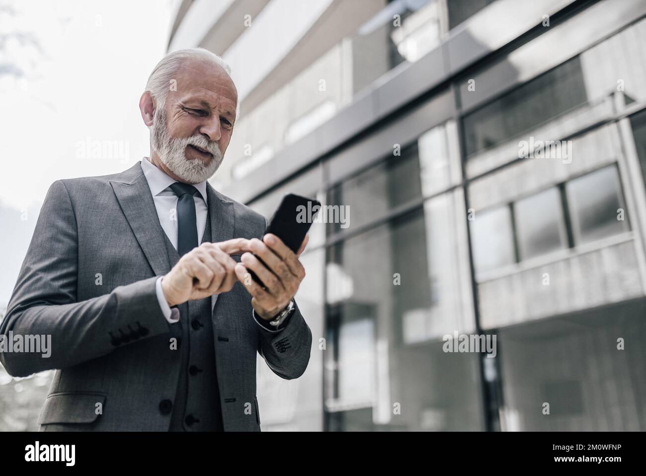 Messagerie professionnelle professionnelle souriante sur smartphone. L'entrepreneur âgé se trouve à l'extérieur de l'immeuble de bureaux. Il porte une combinaison complète. Banque D'Images