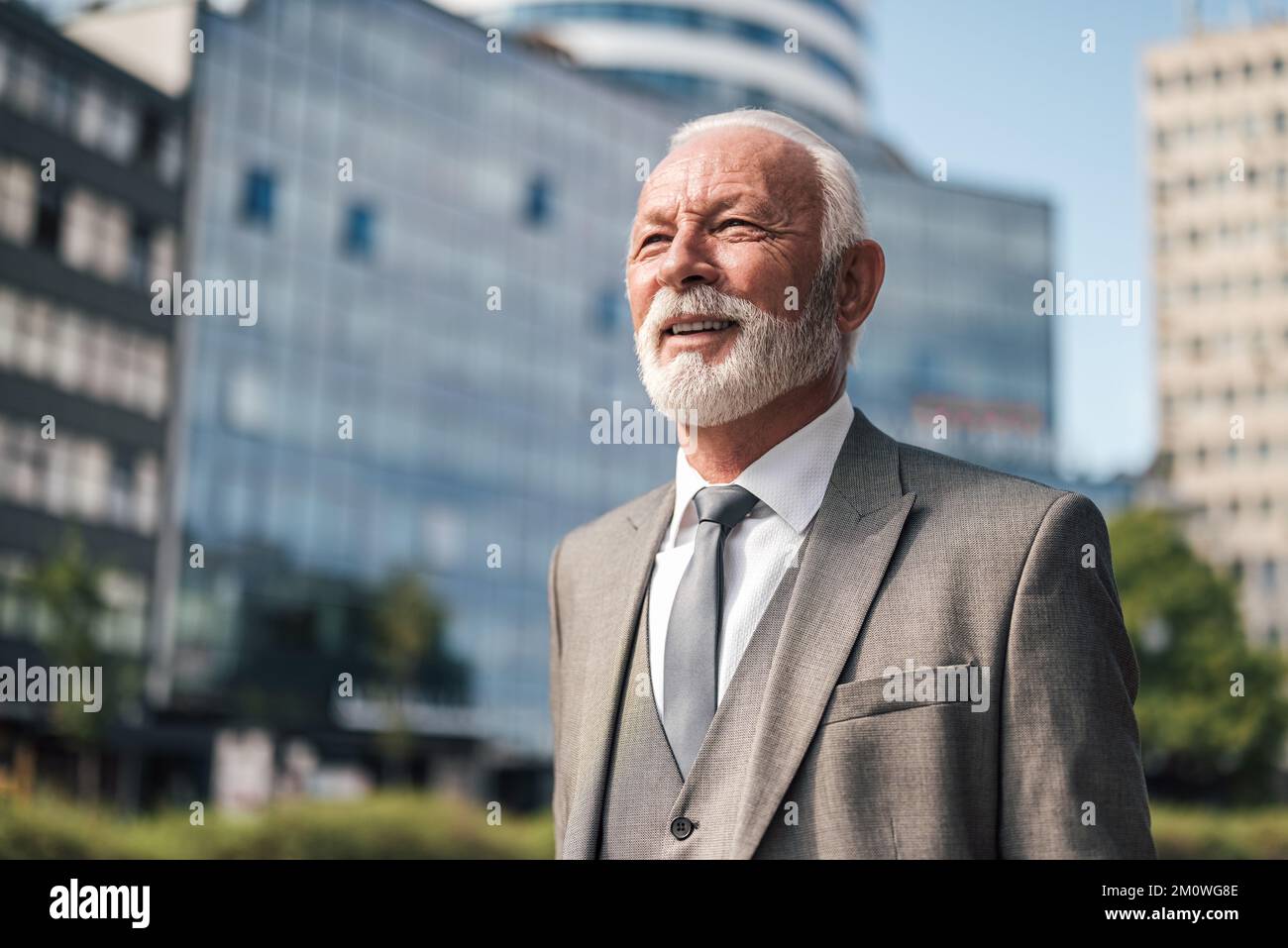 Entrepreneur âgé souriant qui regarde loin tout en se tenant contre les immeubles de bureaux d'affaires Banque D'Images