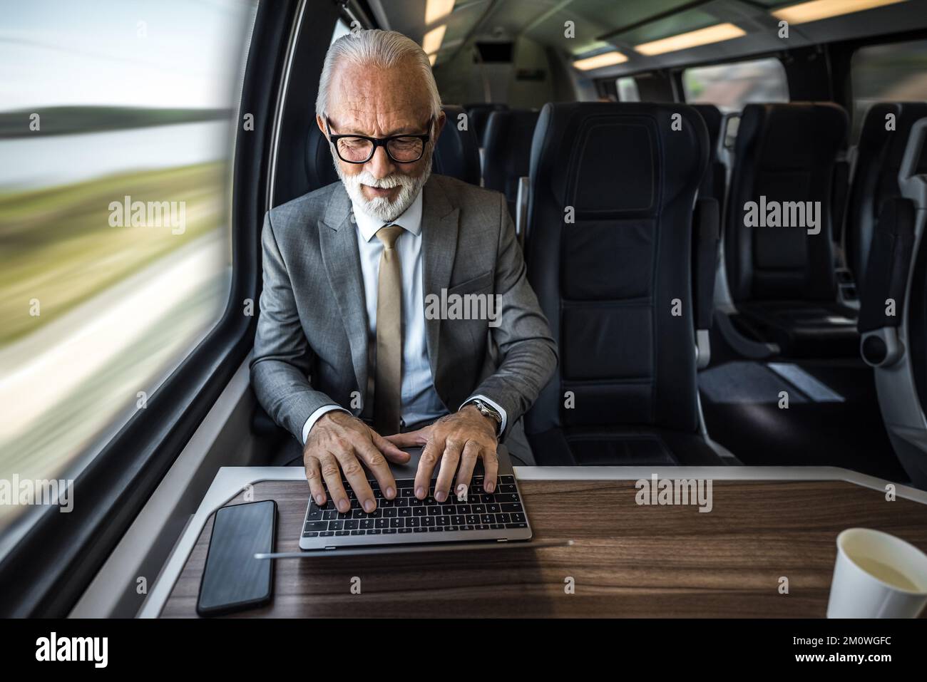 La vue en grand angle d'un homme professionnel utilise un ordinateur portable sur une table. L'homme d'affaires senior est assis par la fenêtre dans le train de voyageurs. Il porte un costume formel d Banque D'Images