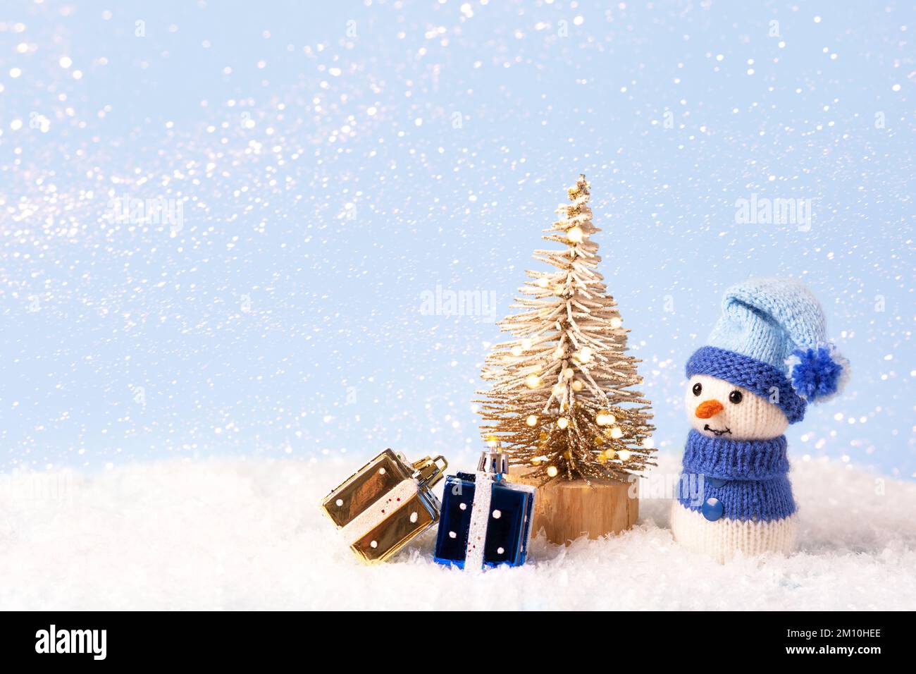 Composition de conte de fées de Noël d'un bonhomme de neige tricoté dans un bonnet et un chandail bleus. Petit arbre de Noël avec deux cadeaux jaunes et bleus Banque D'Images