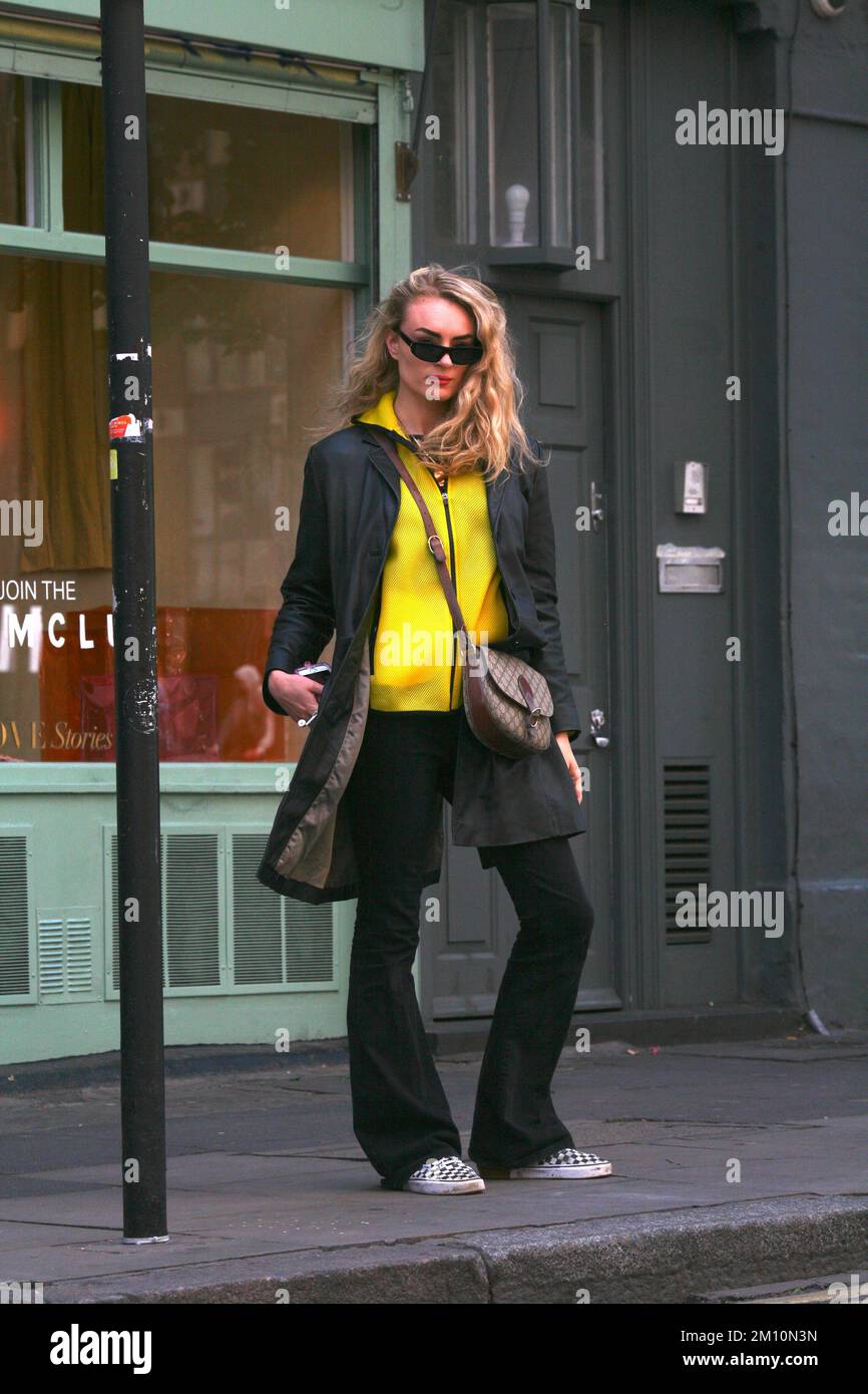 Chic jeune femme style rue, portant long manteau noir, sac à main pantalon noir, baskets, haut jaune et lunettes de soleil sur une rue à Londres Banque D'Images