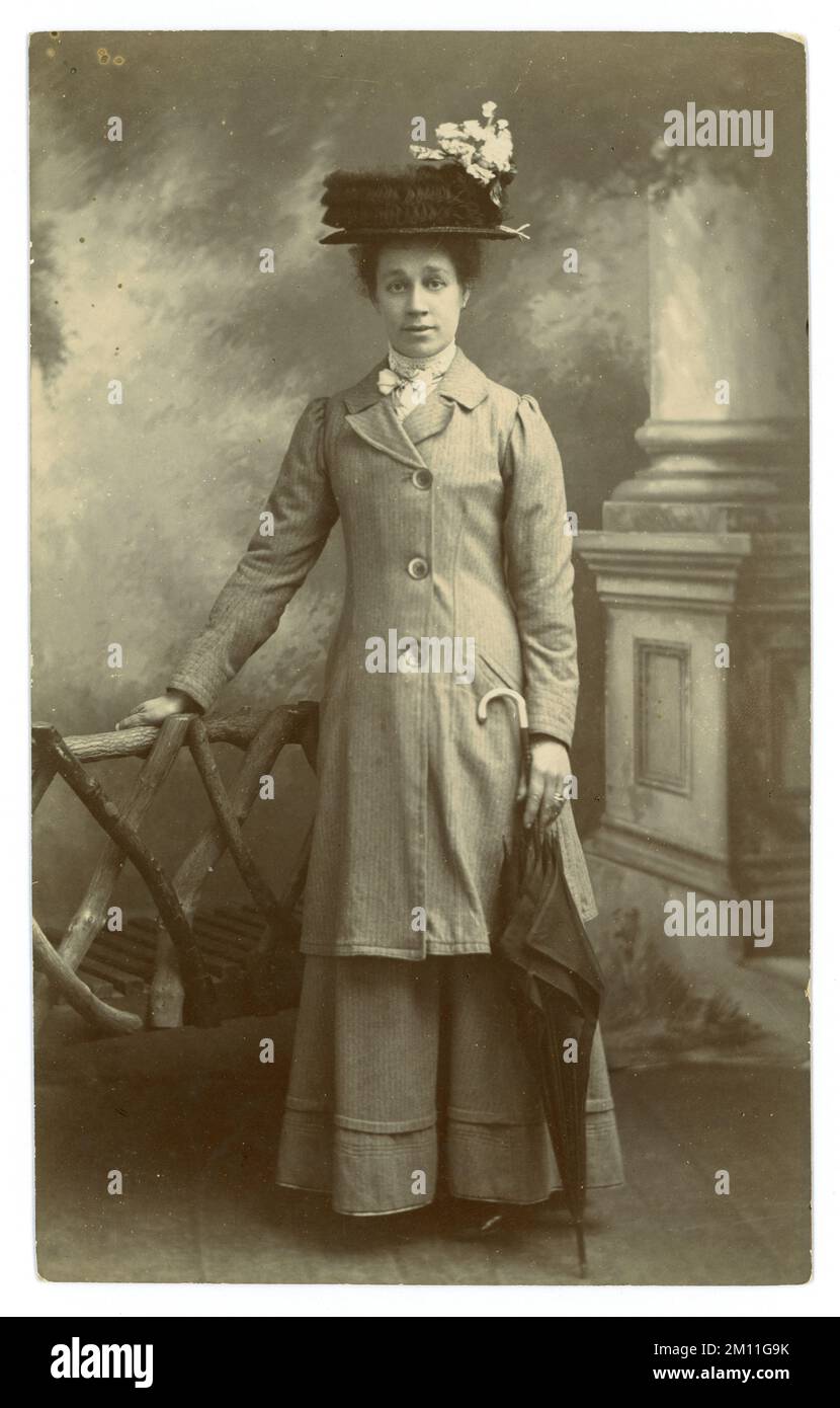 Original Edwardian Era carte postale studio de femme attrayante en veste longue et jupe, grand chapeau avec dentelle ruchée et branche de fleur, tenant un parapluie avec poignée de canne. Dame édouardienne. Femme édouardienne. Daté de juillet 1910, Royaume-Uni Banque D'Images