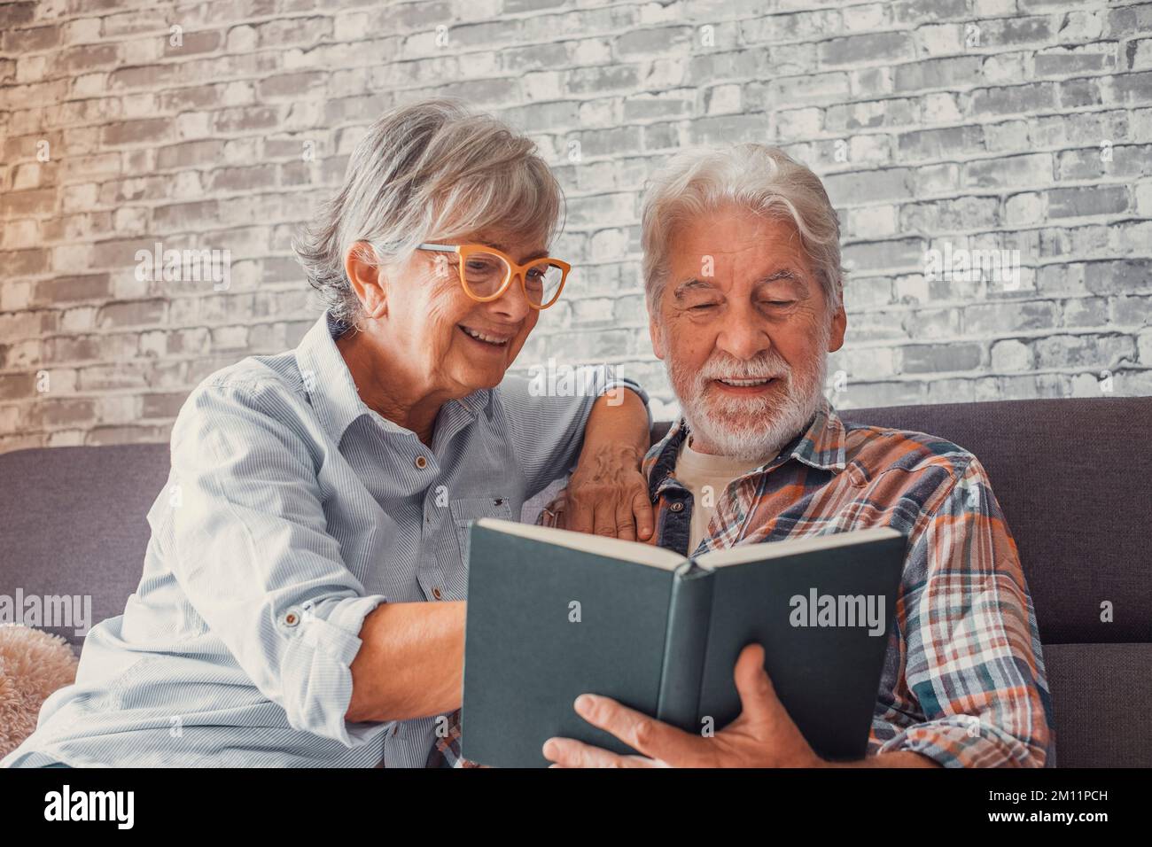 Portrait d'un couple de deux adorables grands-parents lisant un livre ensemble assis sur le canapé. Grand-mère et grand-père week-end de détente à temps libre. Banque D'Images